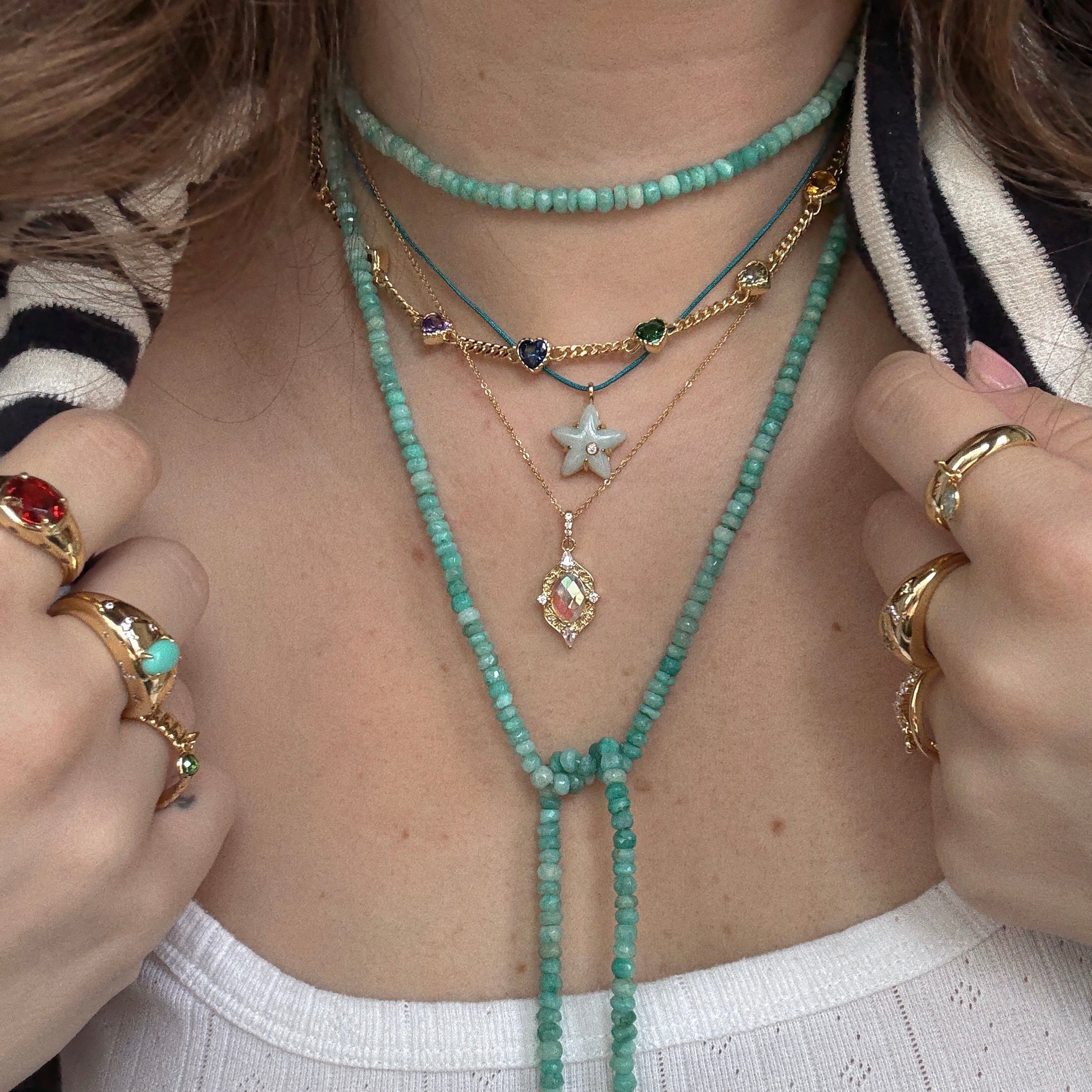 A person wearing layered turquoise bead necklaces, gold chains with gemstone charms, the Turquoise Sky Dome Ring, and gold rings with colorful stones, dressed in a white top and striped jacket.