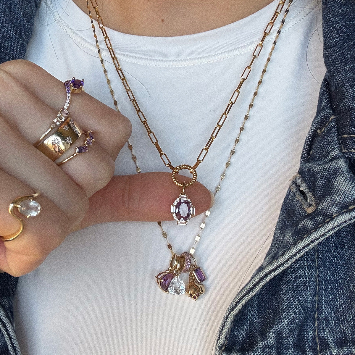 A person in a white top and denim jacket showcases layered gold necklaces, including the Solid Gold Vintage Inspired Birthstone Charm, along with gold rings featuring clear stones and vintage-inspired jewelry with purple gemstones.