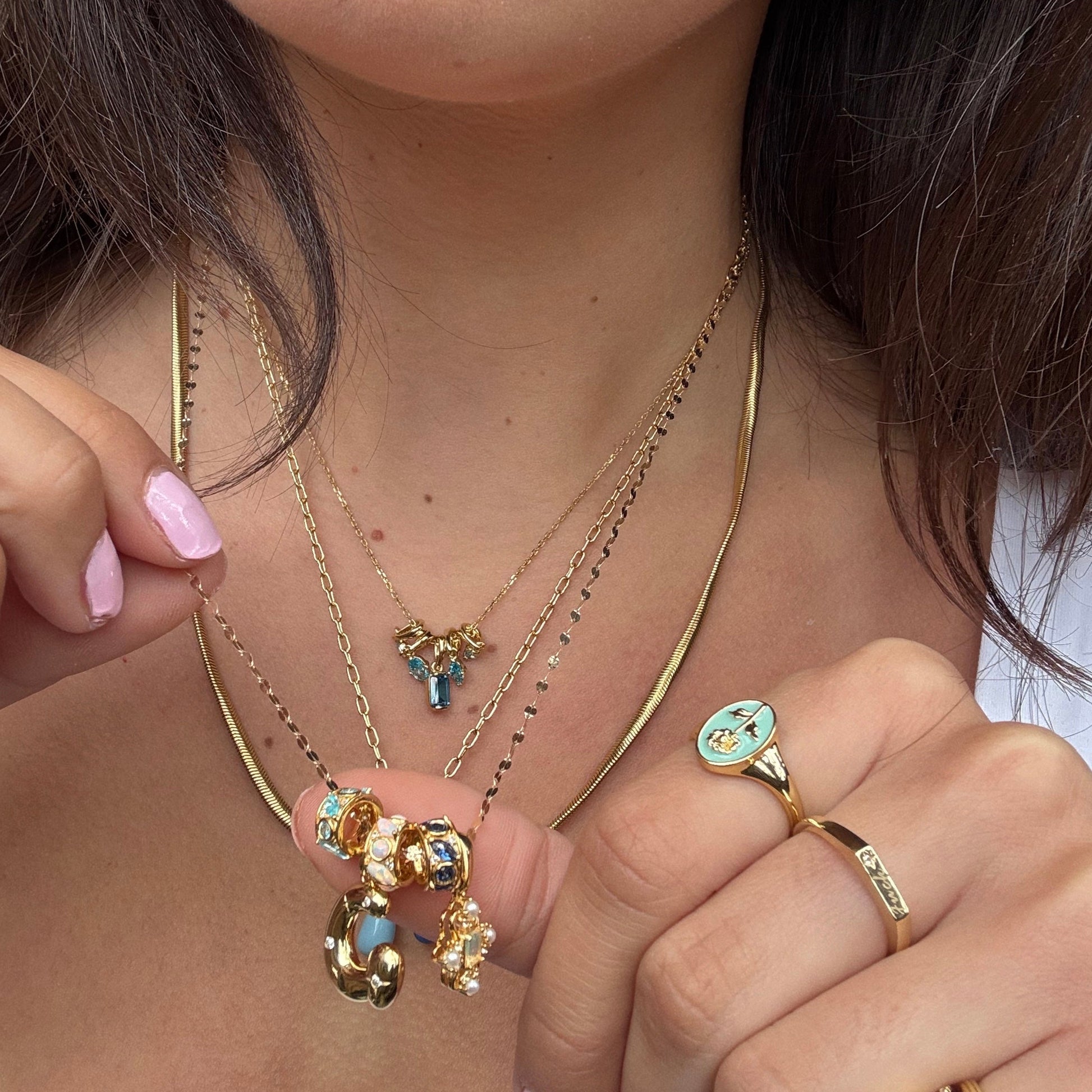 A woman with light pink nail polish wears layered gold necklaces featuring the Shades of You Birthstone Spacer Charm, colorful gold-plated pendants, and a gold ring with a turquoise accent and small face design, holding the necklaces to her neck.