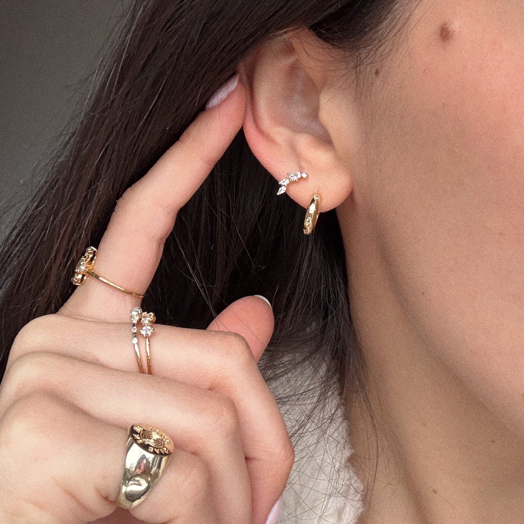 A close-up of a woman’s ear wearing the Classic Birthstone Earring Set—14k gold plated small hoops and a stud earring. Her hand, with gold rings, touches her earlobe. She has light skin and long brown hair.
