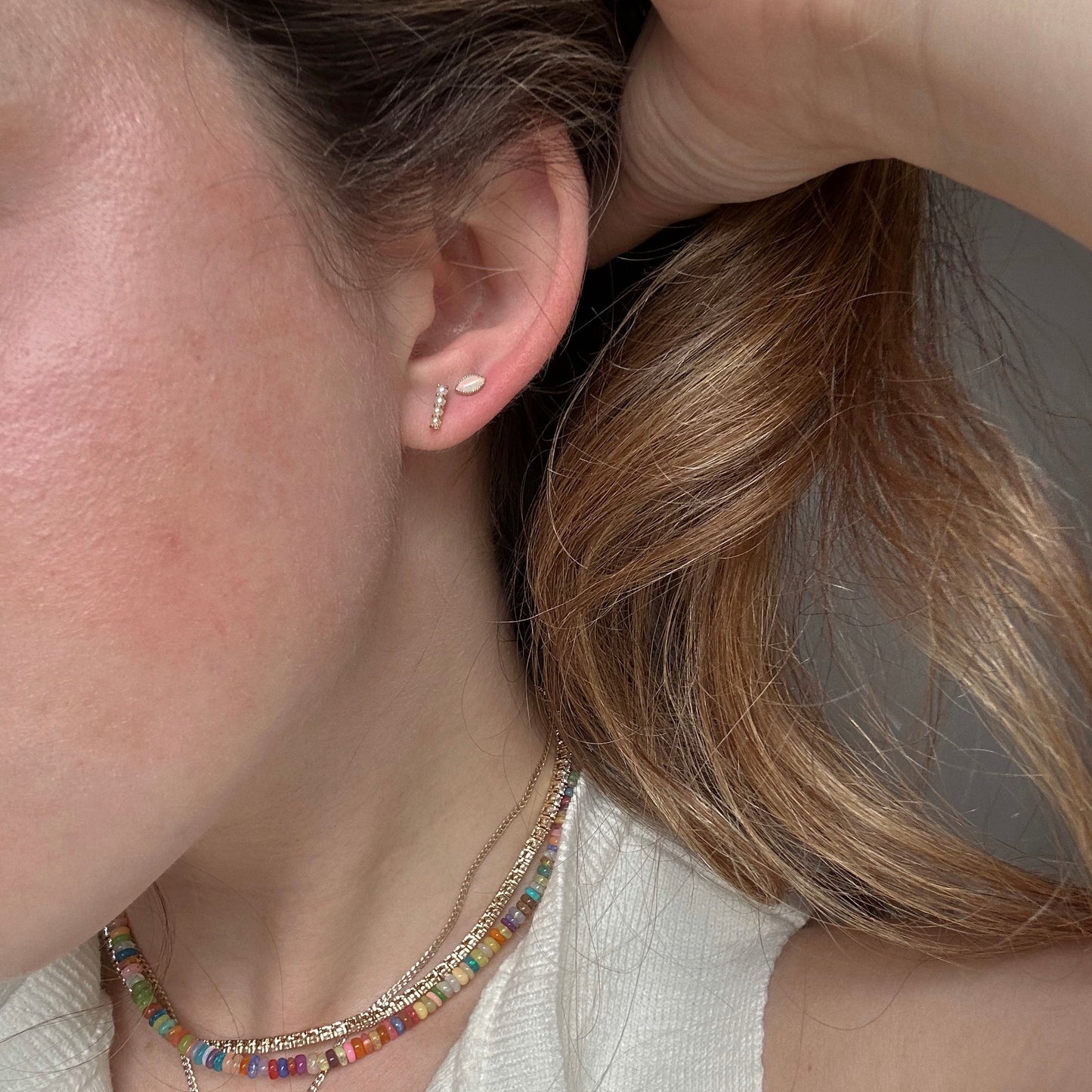 A close-up of a person’s ear shows two Solid Gold Opal Marquise Flat Back Studs. The person has light brown hair, wears a colorful beaded necklace and a white top, and holds their hair back to display the earrings.