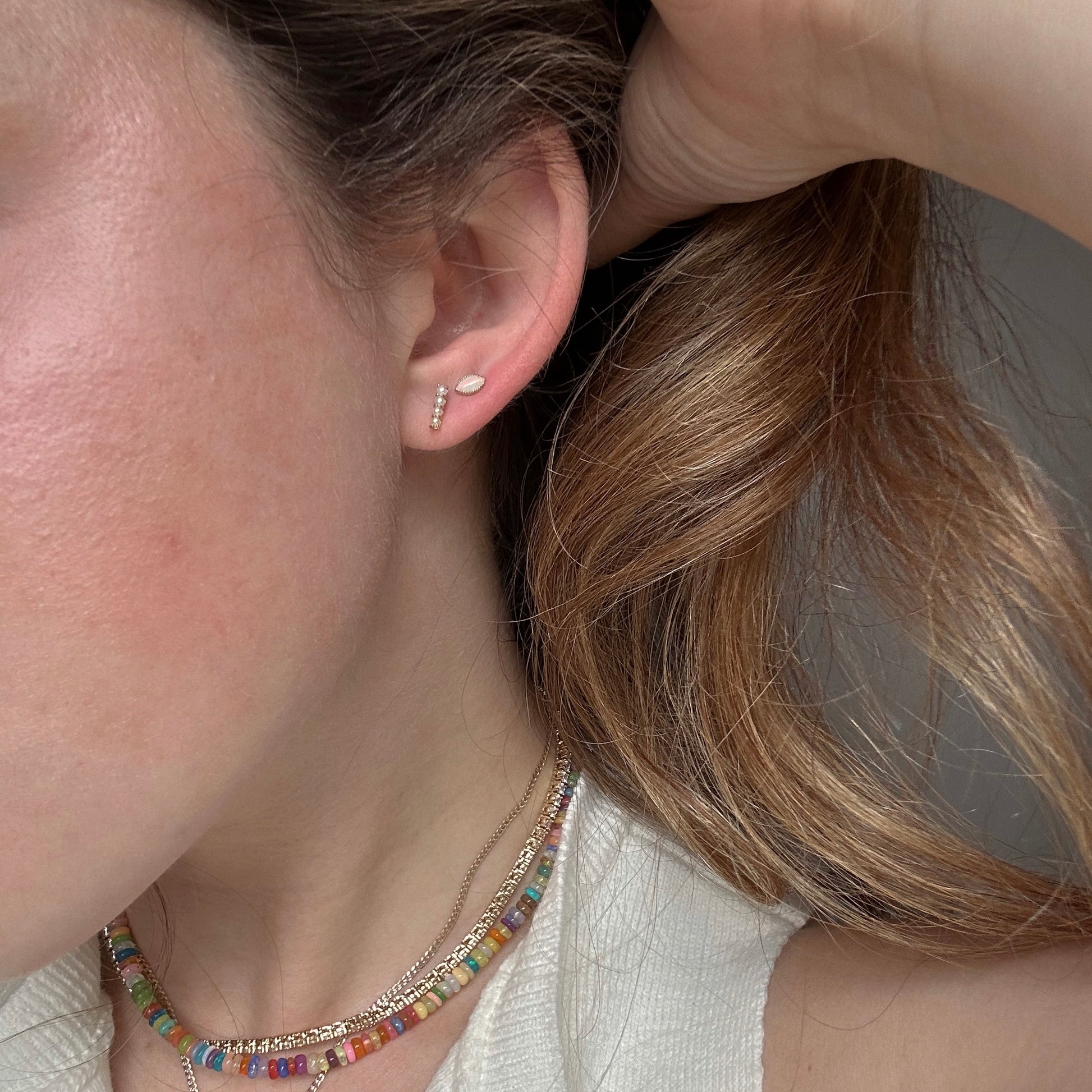 A close-up of a person’s ear shows two Solid Gold Opal Marquise Flat Back Studs. The person has light brown hair, wears a colorful beaded necklace and a white top, and holds their hair back to display the earrings.