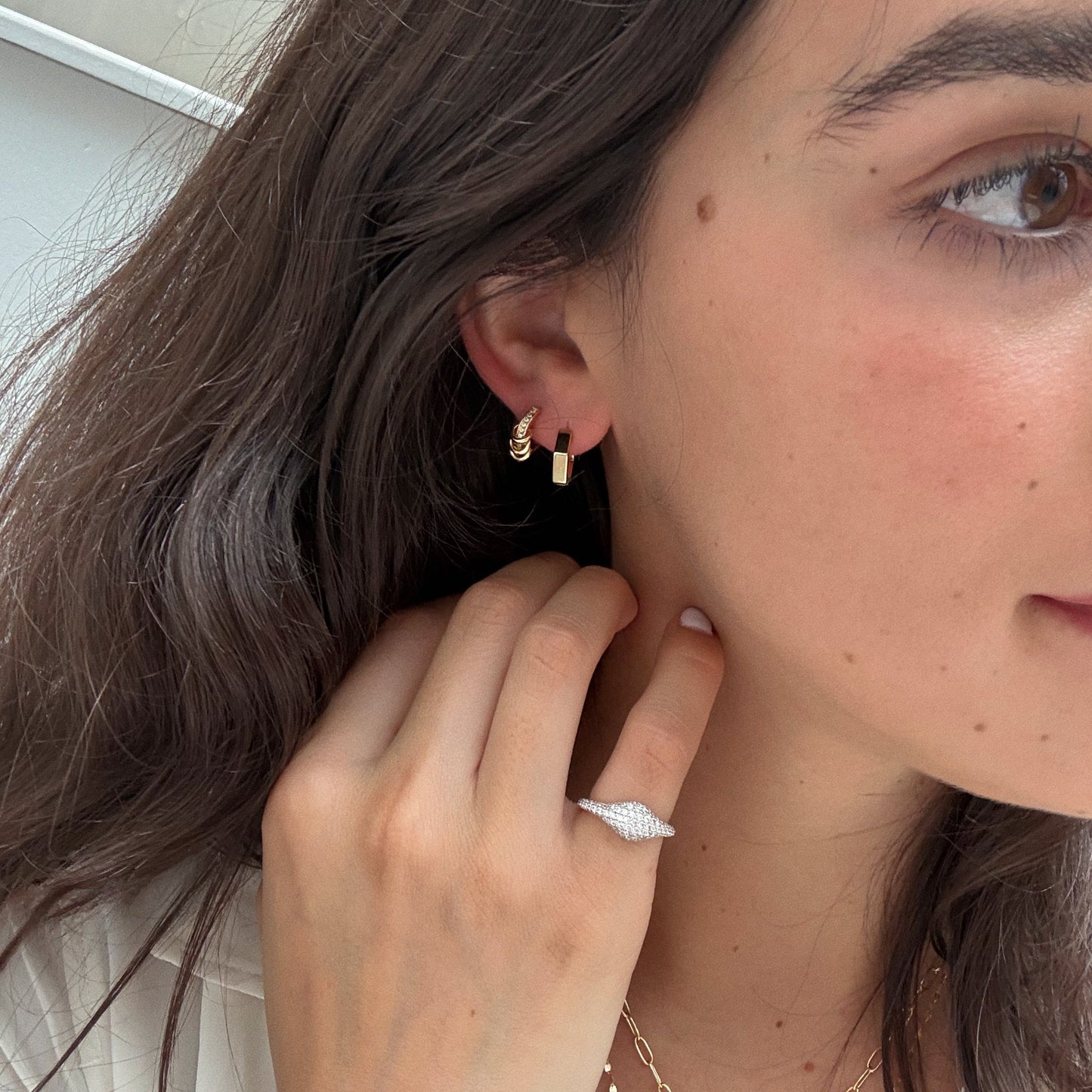 A close-up of a woman’s face and hand showcases Levi Huggies in 18k yellow gold, a textured white ring, and part of a gold necklace. She has light skin, natural makeup, and wavy brown hair.