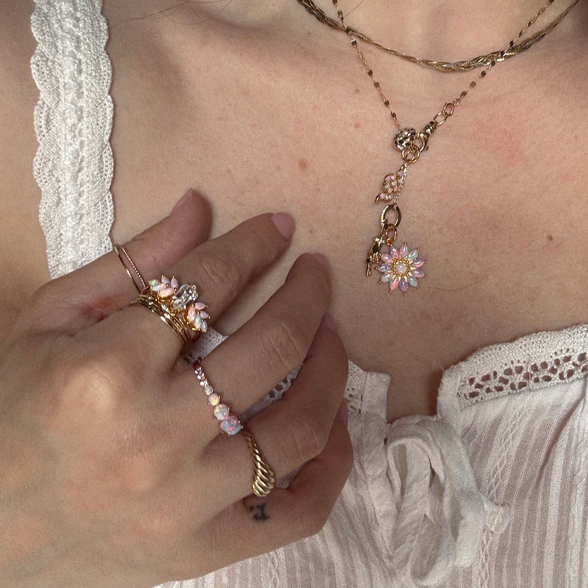 A person wearing a white textured top and layered 18k gold vermeil jewelry, including necklaces, bracelets with pink and white flower charms, and the Pink Opal Ombre and Topaz Lotus Ring. Their hand gently touches their collarbone.
