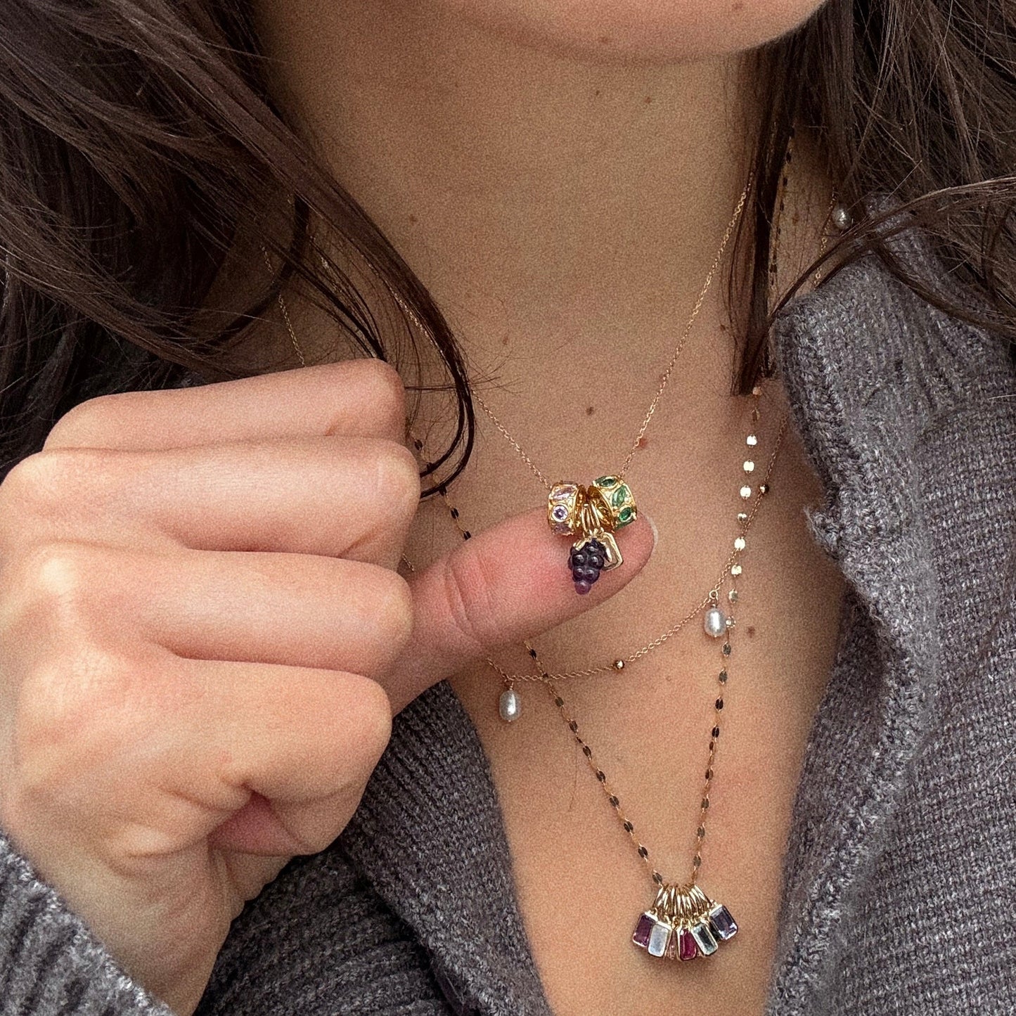 A person in a gray knit top wears layered gold necklaces, including the Solid Gold Foodie Charm with tiny, vibrant fruit-shaped charms, and holds up a pendant adorned with colorful fruit designs.