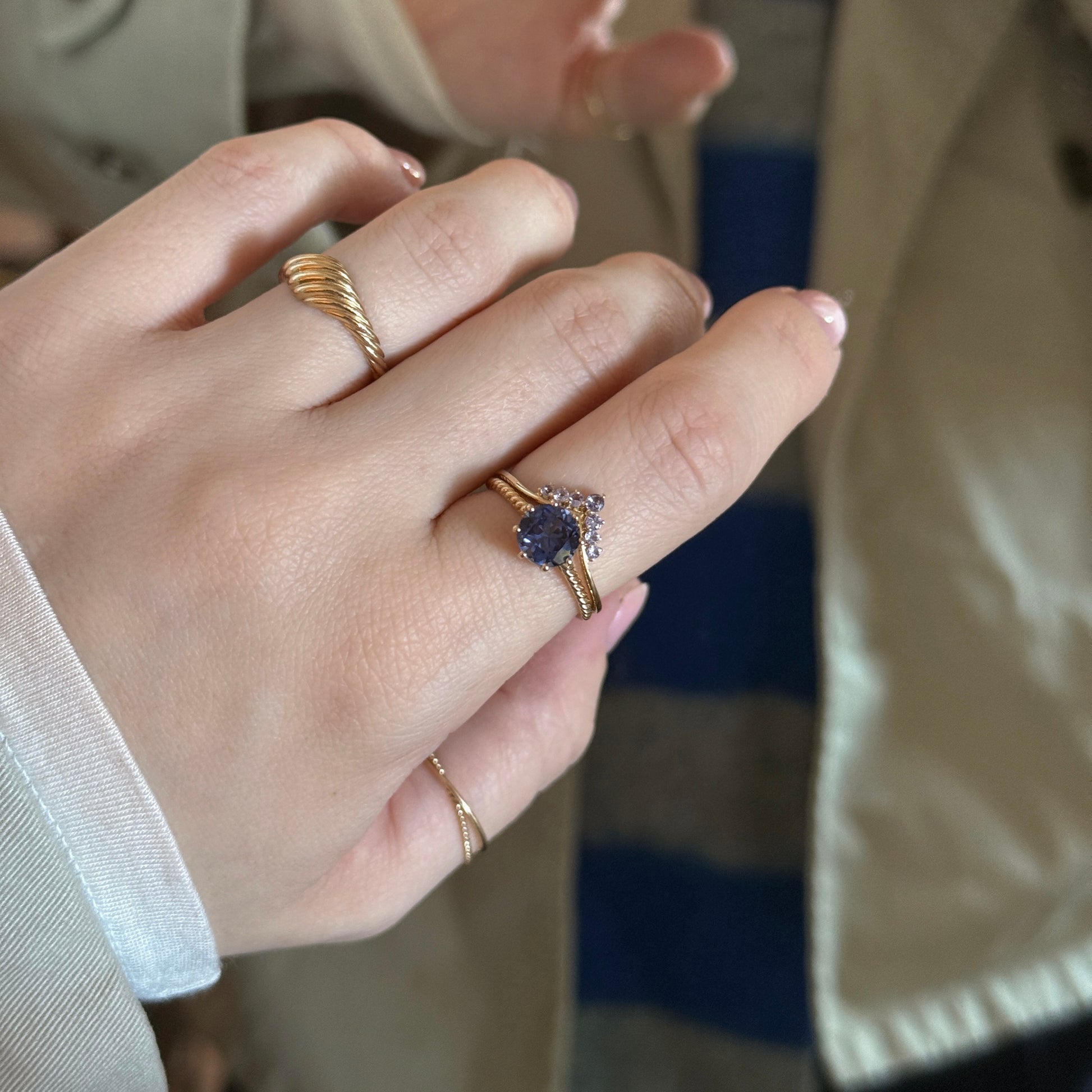 A hand with neatly manicured nails wears the Solid Gold 2025 December Capsule Tanzanite Ring Set (with Free Gift), featuring a purple gemstone, in front of someone dressed in a beige coat and blue-striped garment.