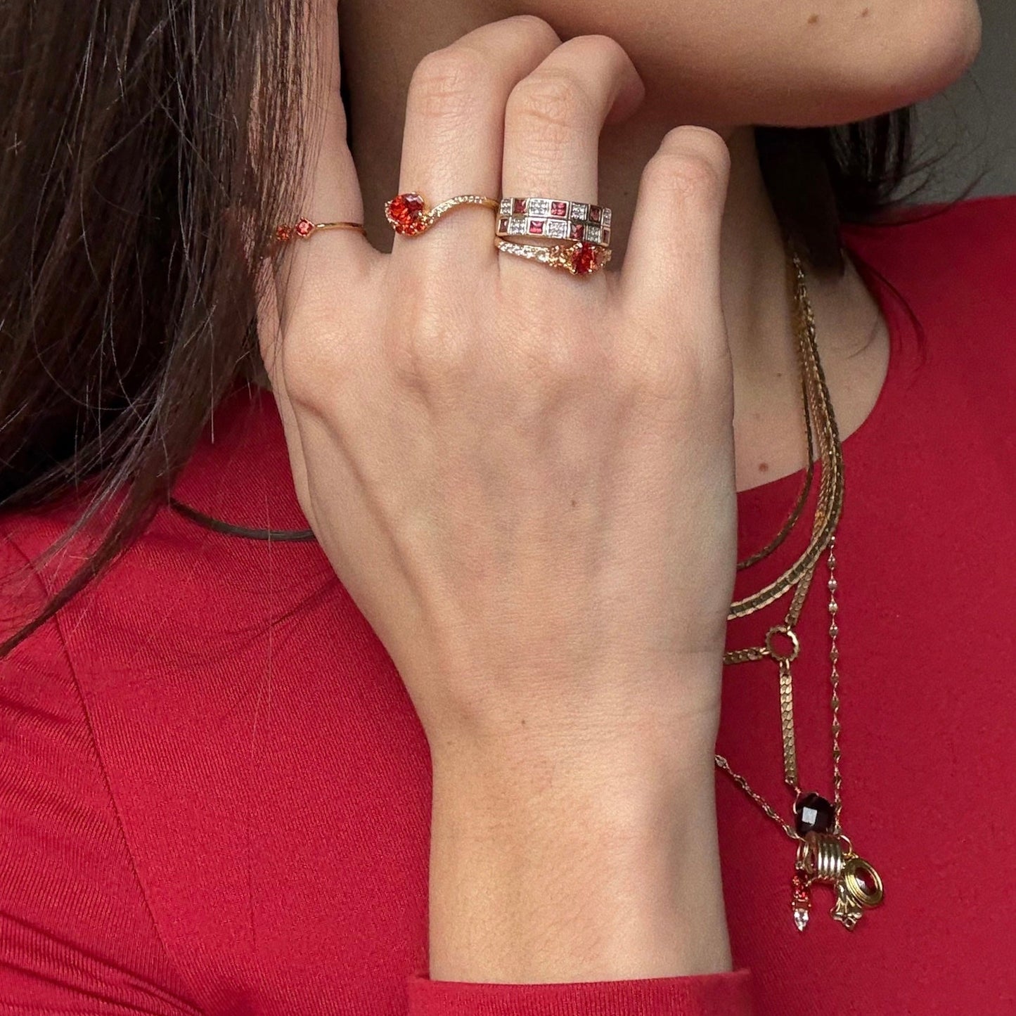 A woman in a red top showcases her hand wearing the Main Character Floating Ombre Birthstone Ring with red and clear stones, paired with layered gold necklaces featuring assorted pendants and a bold red gemstone.