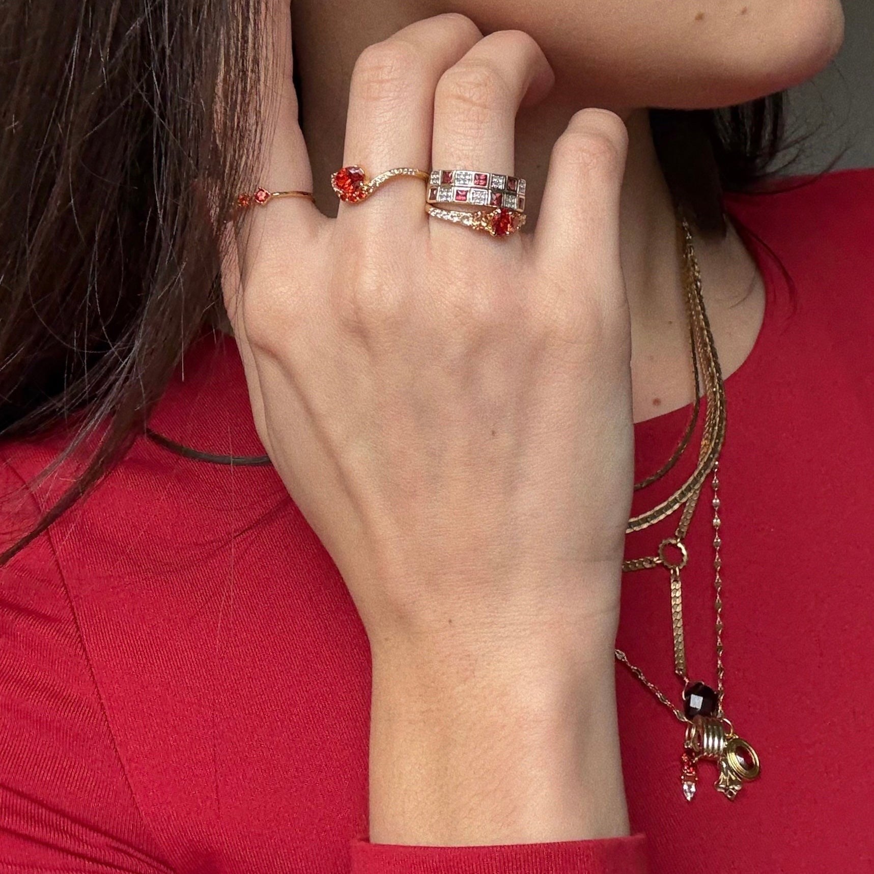 A woman in a red top showcases her hand wearing the Main Character Floating Ombre Birthstone Ring with red and clear stones, paired with layered gold necklaces featuring assorted pendants and a bold red gemstone.