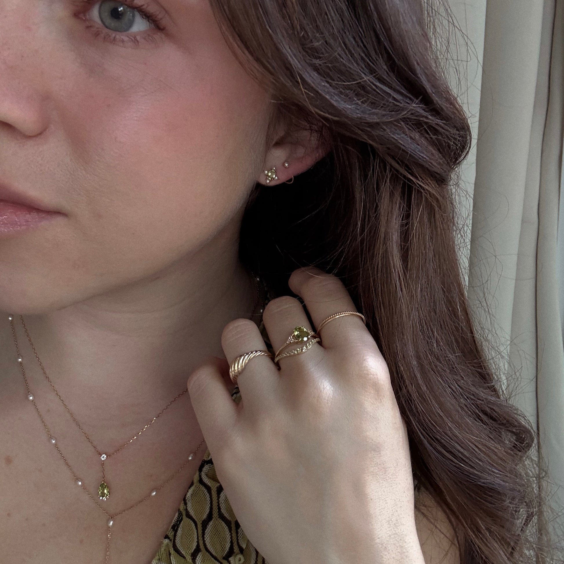 A woman with long brown hair wears gold rings, necklaces, and Solid Gold 2025 August Capsule Vintage Inspired Peridot Stud Earrings. Her hand is near her face, accenting the peridot jewelry. She has light skin, a patterned top, and a curtain behind her.