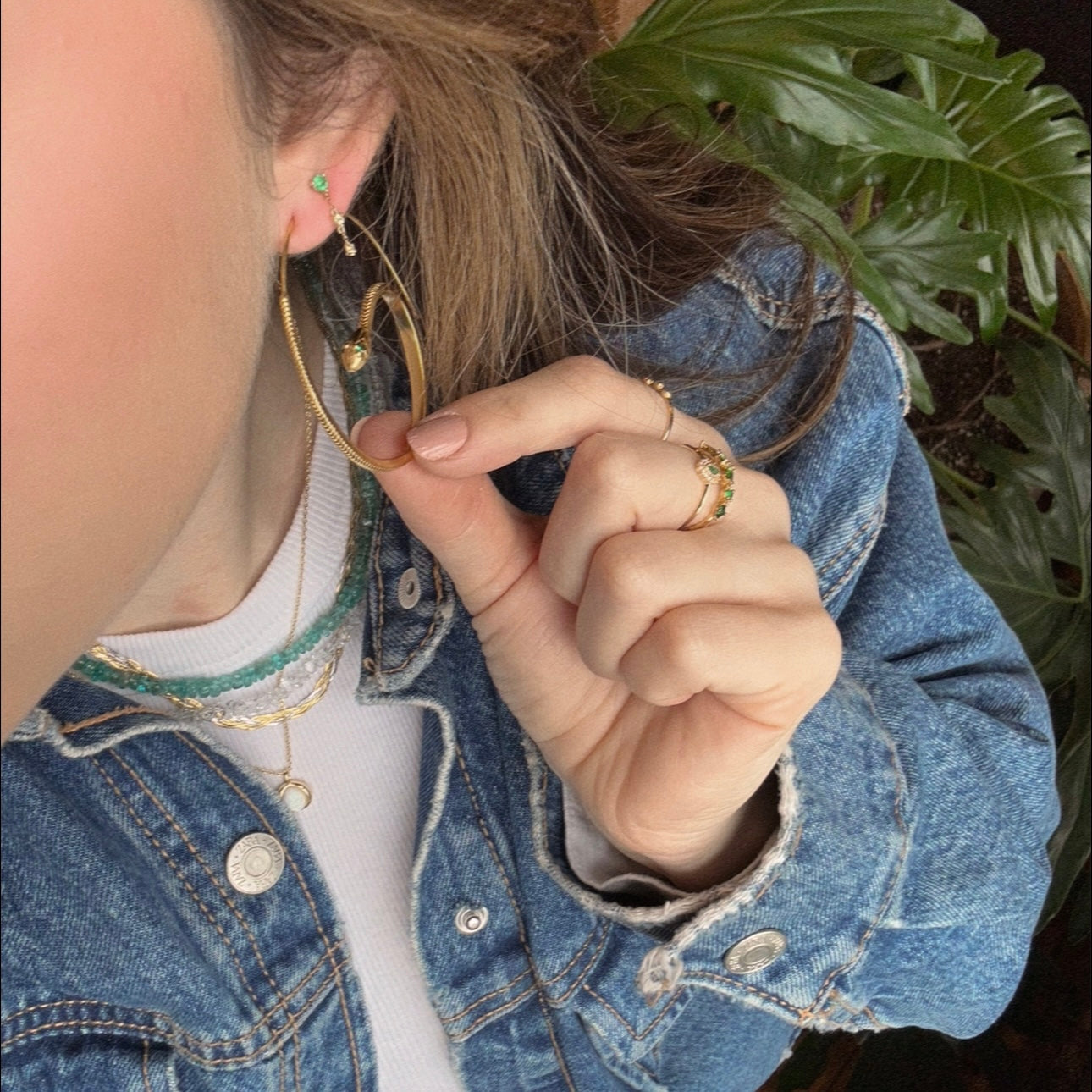 A person in a denim jacket holds the Snake Hoop Earrings, plated in 18k yellow gold. They wear layered necklaces and rings, such as a turquoise beaded necklace and a gold ring with green stones. A leafy plant is in the background.