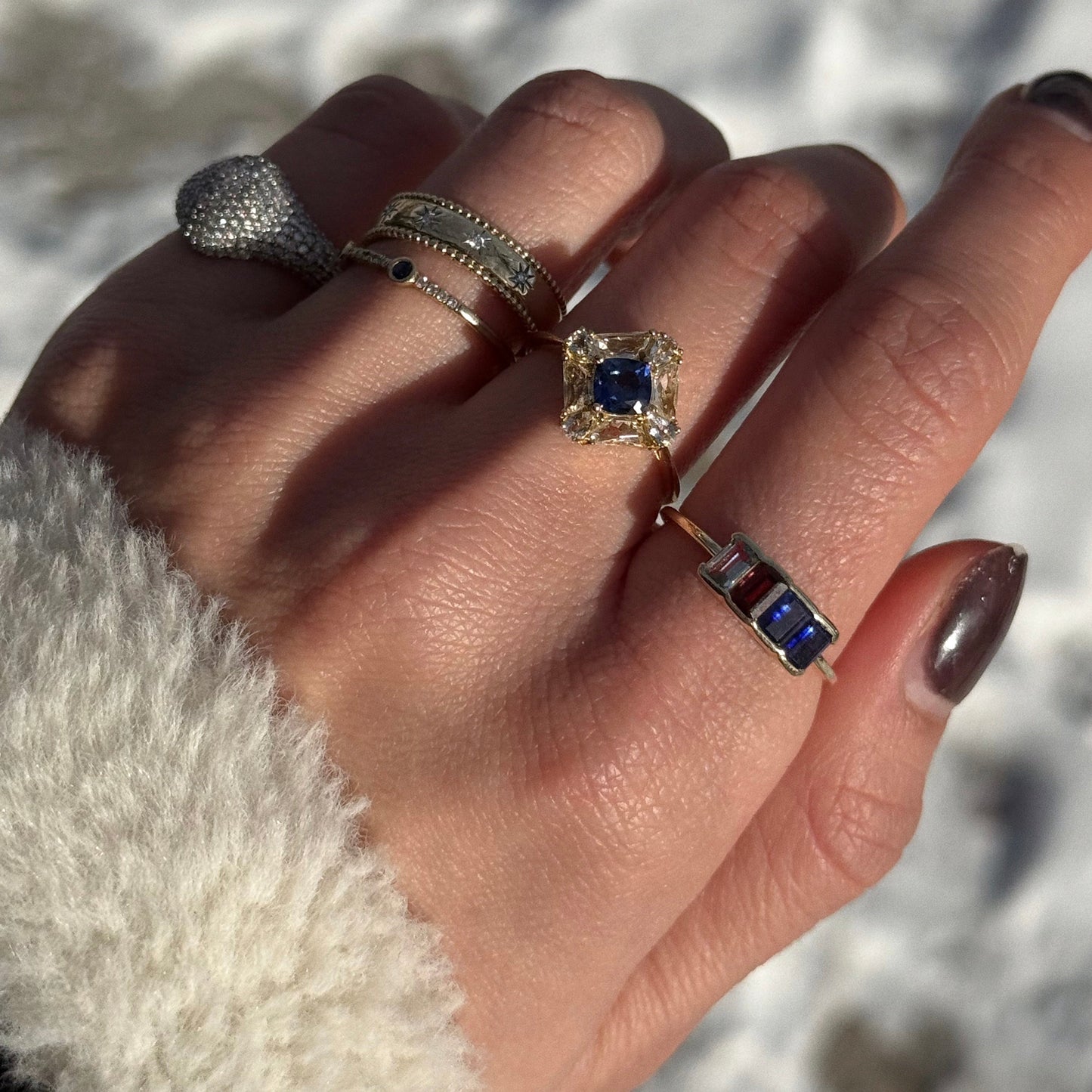 A hand with four rings—a diamond-studded dome, a thin gold band with tiny gems, a square blue gemstone ring, and a Solid Gold Aquamarine Sapphire and Garnet Baguette Gemstone Ring—shown against a snowy background.