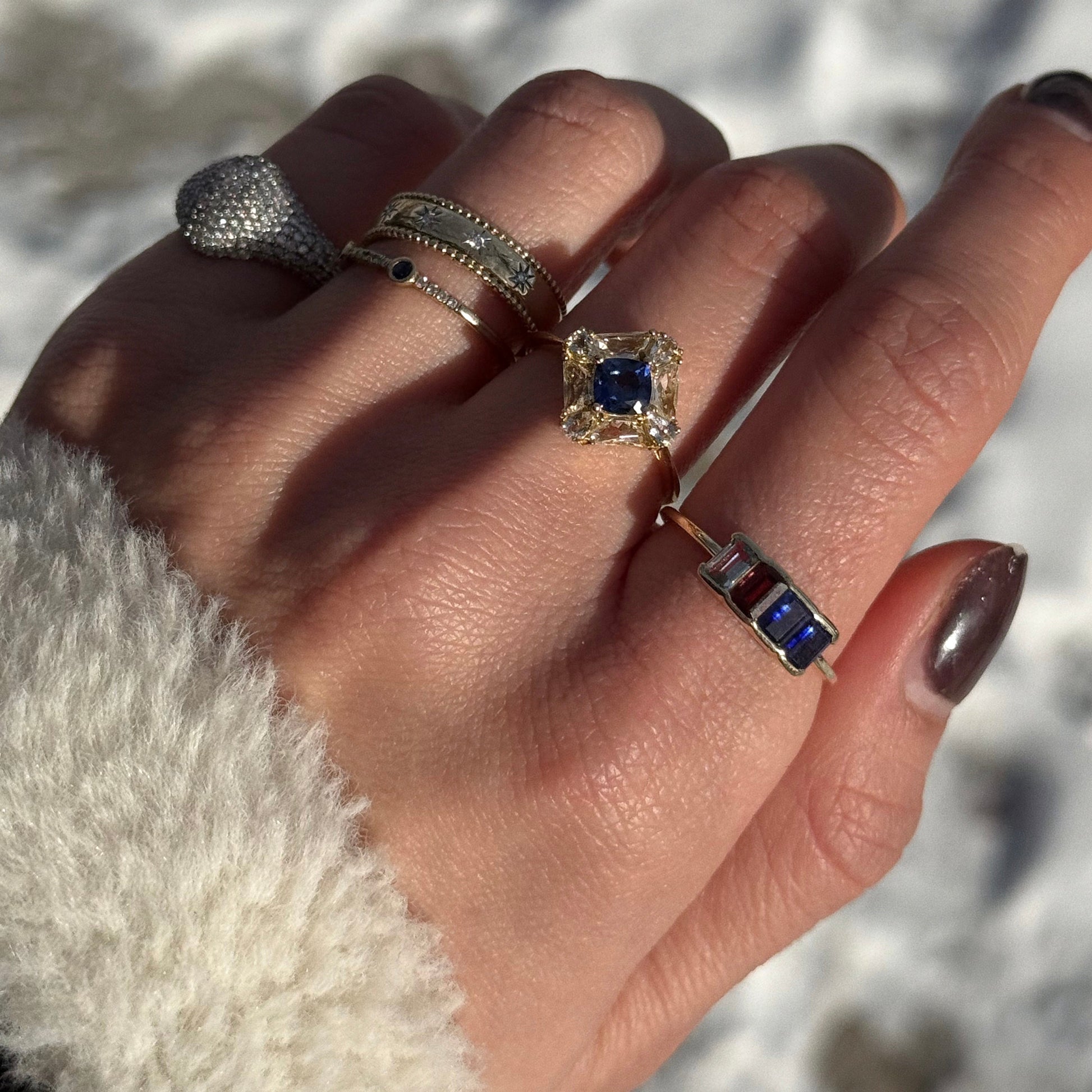A hand with four rings—a diamond-studded dome, a thin gold band with tiny gems, a square blue gemstone ring, and a Solid Gold Aquamarine Sapphire and Garnet Baguette Gemstone Ring—shown against a snowy background.