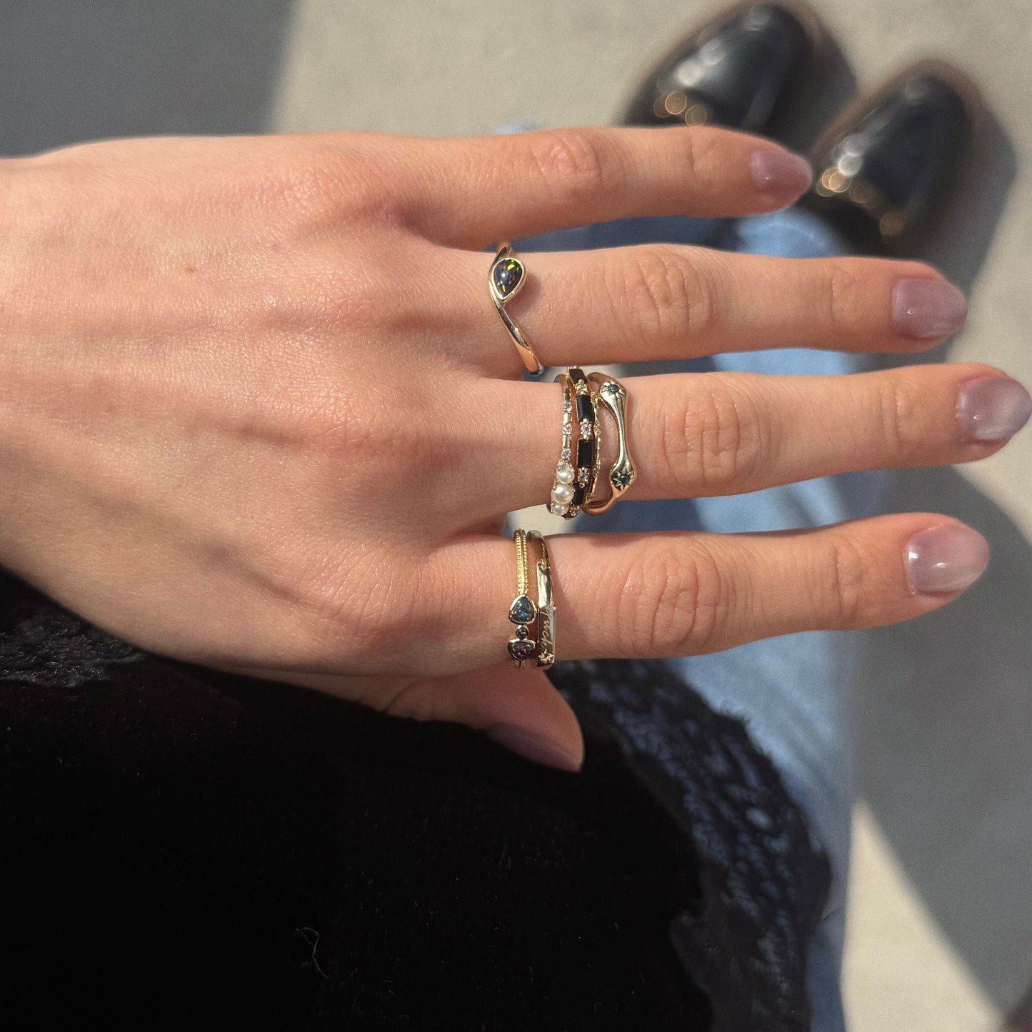 A hand with neatly manicured nails wears the Solid Gold Black Opal Stars Aligned Ring and yellow gold bands, all resting on denim-clad legs. The background includes black shoes and a shadowed surface.
