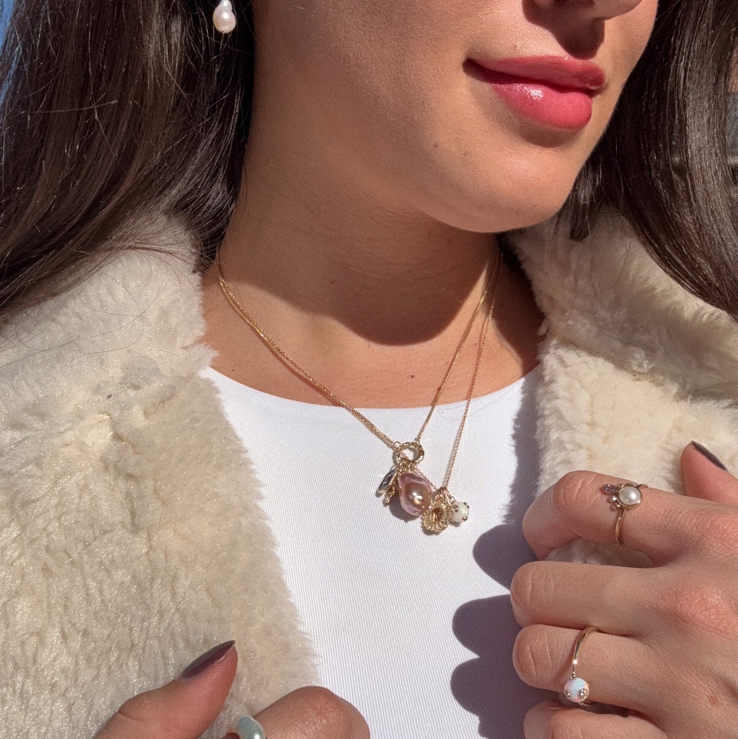 A woman in a white top and cream fur coat showcases pearl jewelry—a gold necklace with a large pearl pendant, a pearl earring, and the Solid Gold Gem Drop Opal Spinner Ring—paired with light pink lipstick and manicured nails.