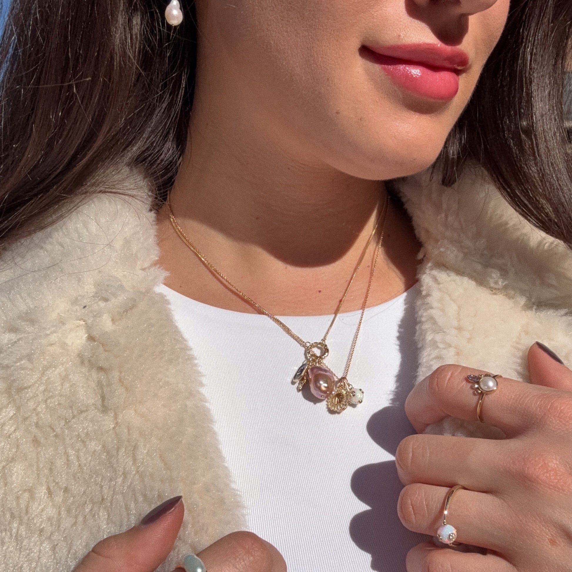 A woman in a white top and cream fur coat showcases pearl jewelry—a gold necklace with a large pearl pendant, a pearl earring, and the Solid Gold Gem Drop Opal Spinner Ring—paired with light pink lipstick and manicured nails.