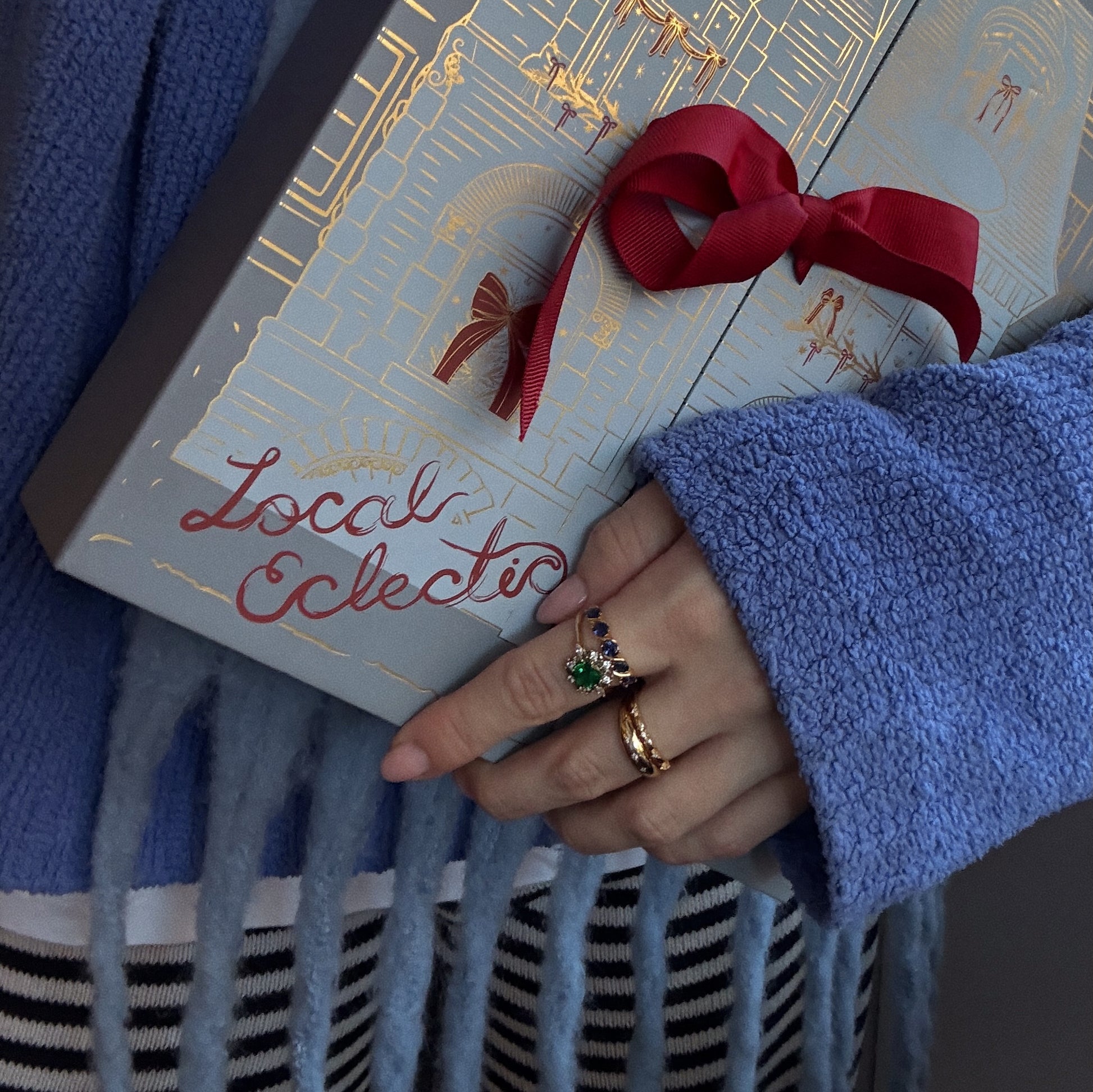 A person in a blue sweater holds a "2025 Jewelry Advent Calendar" box with gold designs and a red ribbon—ideal for holiday gifting. Their hand, adorned with rings, shows stripes peeking from below.
