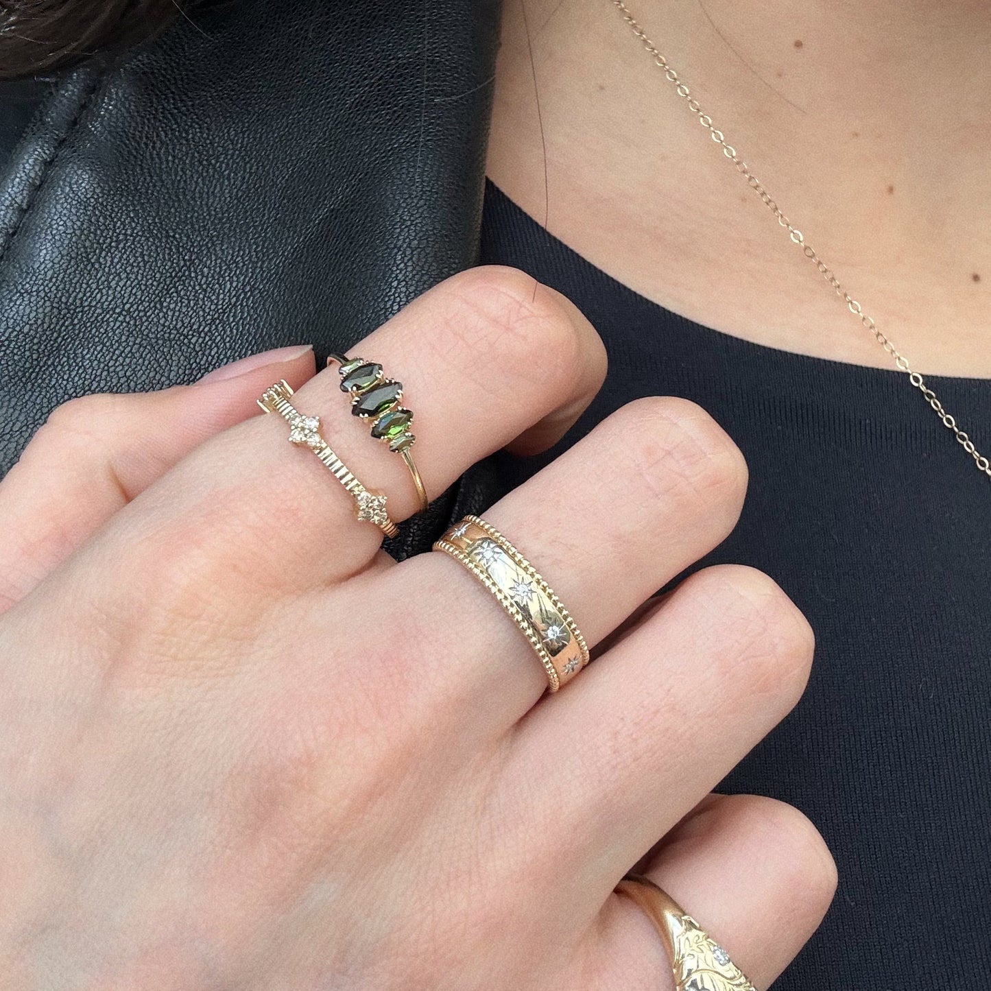 A close-up of a hand wearing the Solid Gold Essential Marquise Cluster Ring, featuring heirloom-quality gold with green tourmaline and small diamonds, shown against a black top and leather jacket. A delicate gold necklace is also visible.
