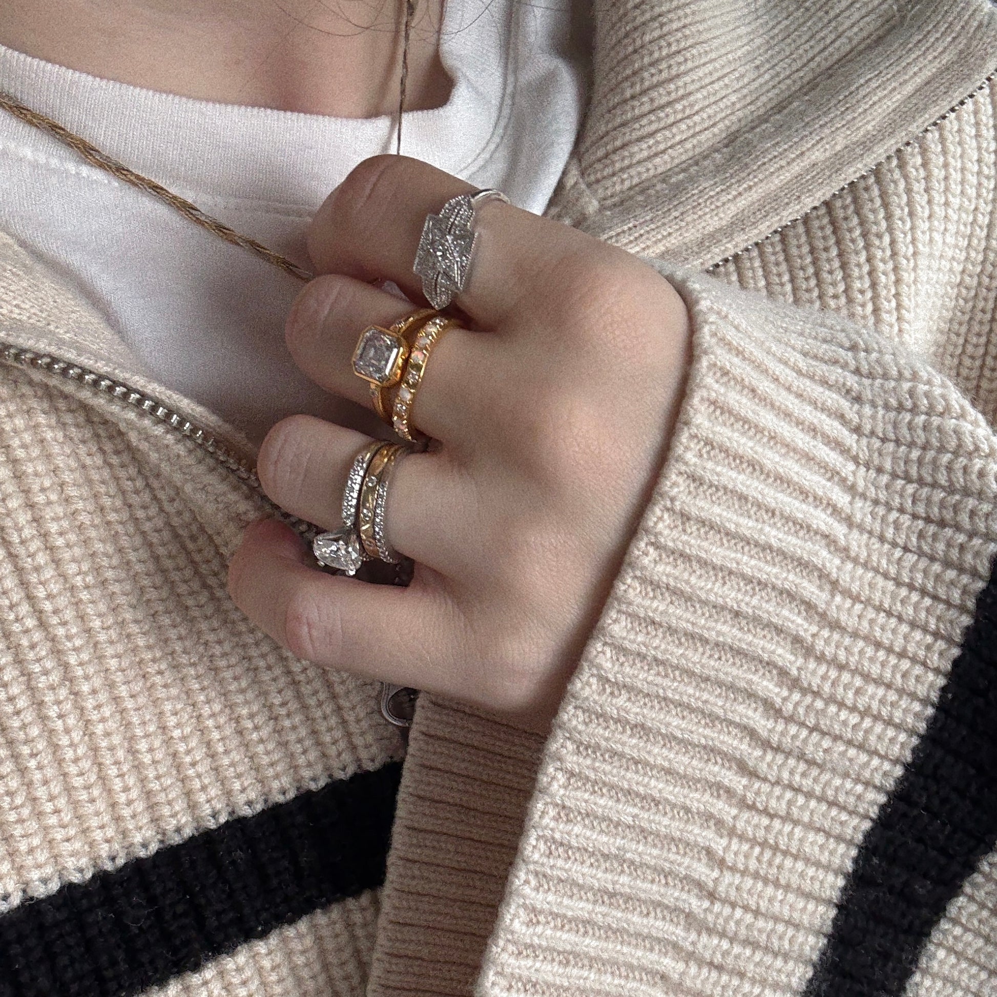 A hand wearing the Art Deco Inspired Green Amethyst Statement Ring is partially covered by the sleeve of a beige knit sweater with a black stripe, layered over a white shirt.