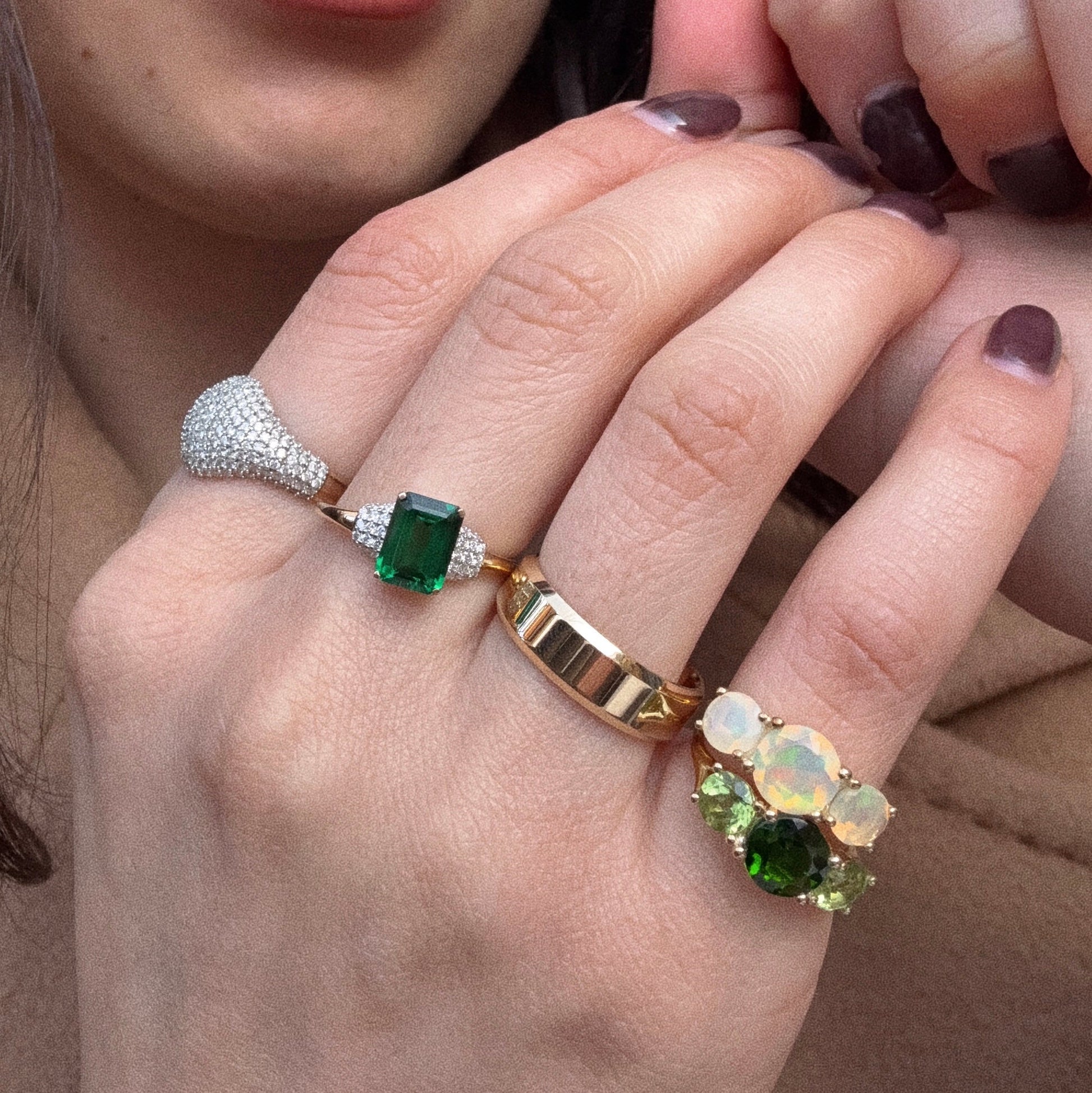 A close-up of a hand with painted dark nails wearing the Solid Gold Trilogy Birthstone Pinky Ring Peridot (Sample Size 4), along with three other gold rings: one plain band, one with opals and green stones, and a diamond-encrusted peridot ring.