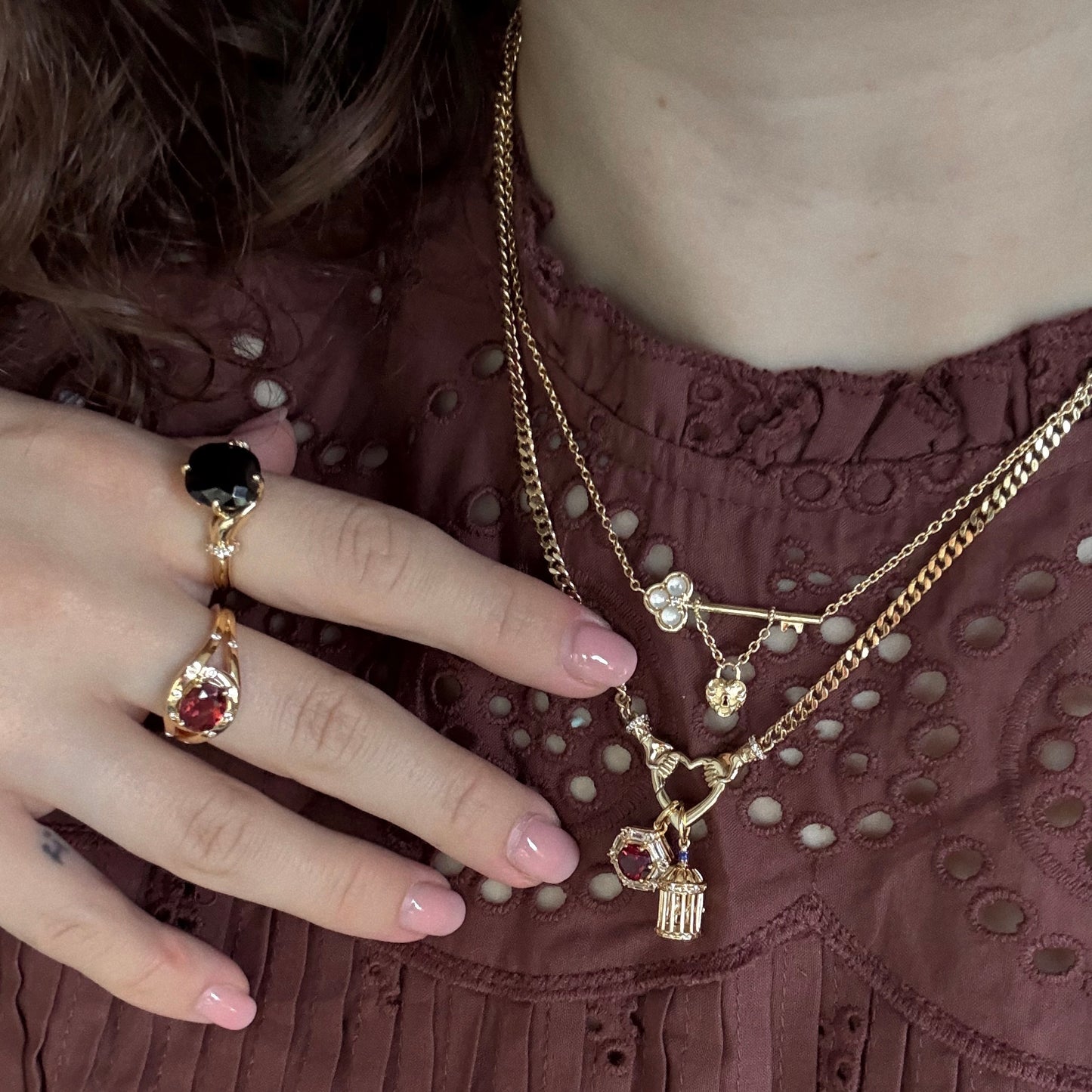 Wearing a maroon eyelet blouse, the person showcases gold jewelry: two layered pendant necklaces and rings, including the Past Life Black Spinel Statement Ring. Their nails are manicured in light pink.
