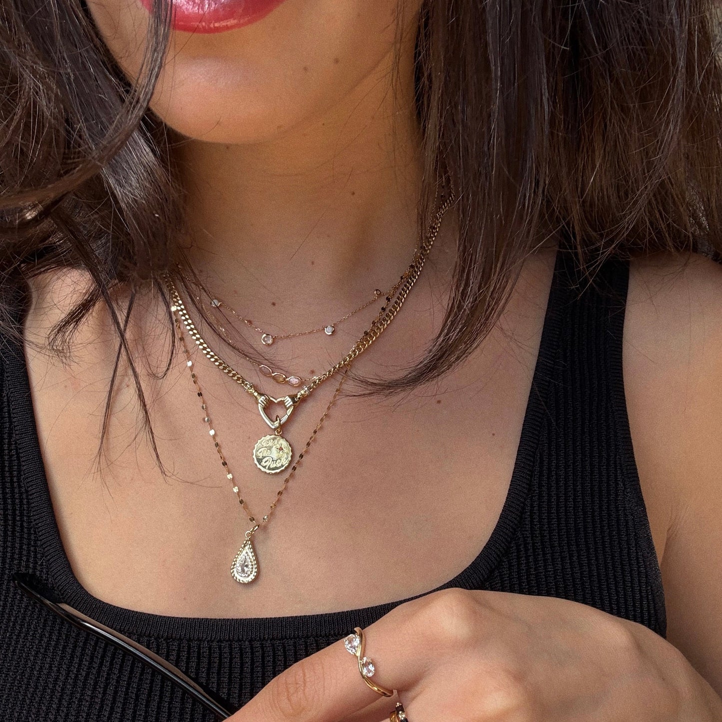 A woman, visible from her lower face to upper torso, wears layered gold necklaces, a black ribbed tank top, and holds sunglasses. She also wears the Solid Gold 2026 April Capsule Floating Duo White Topaz Ring, an elegant limited edition jewelry piece.