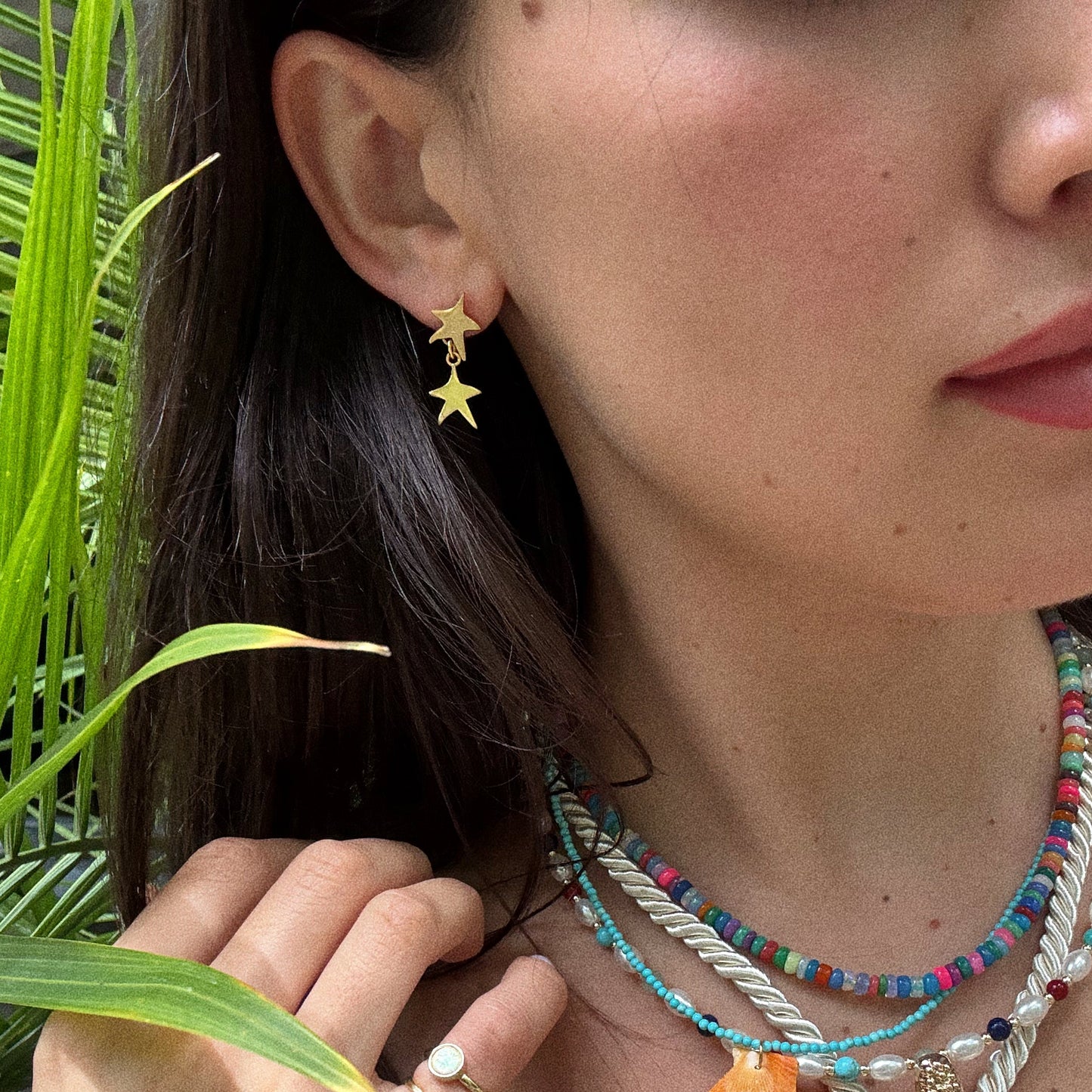 A close-up of a woman in Star Drop Earrings, featuring 18k gold plating, with long dark hair and colorful beaded necklaces beside a green plant.