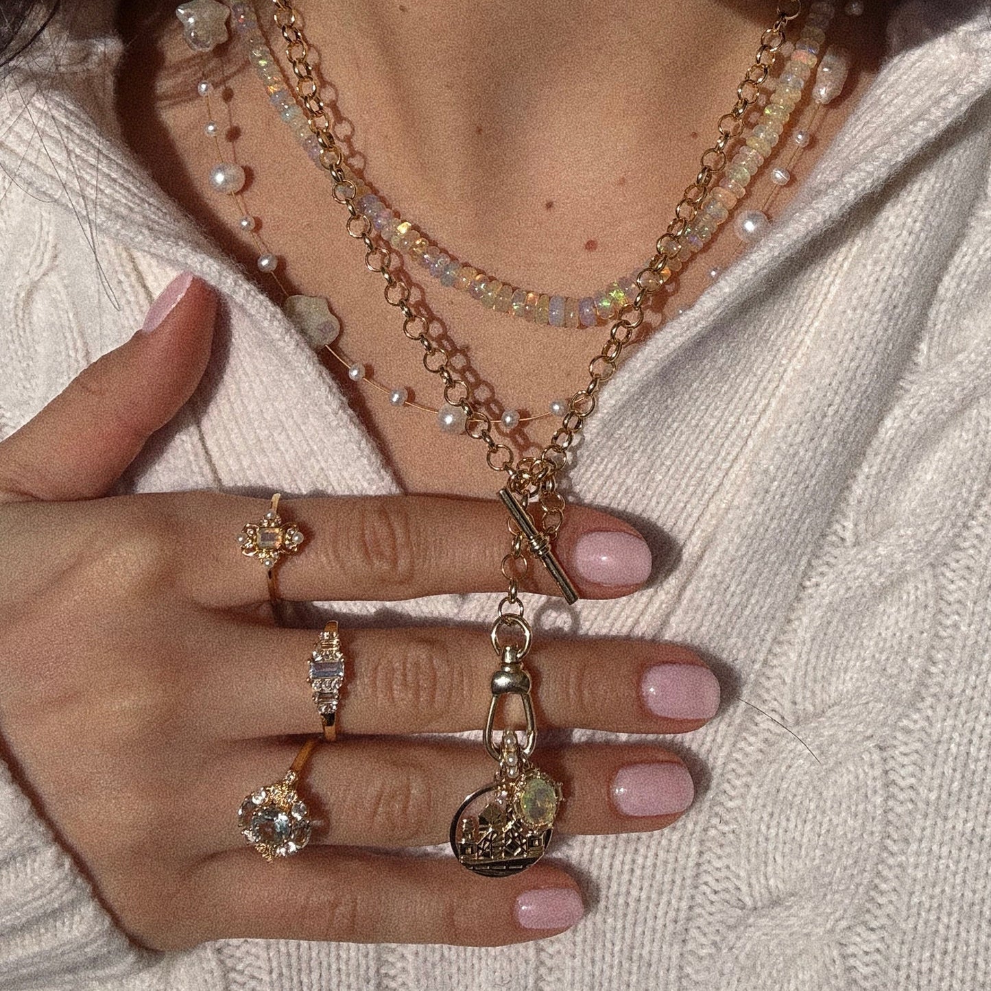 A person with light pink nail polish wears multiple gold rings with gems, a Suki Necklace, and layered gold-plated necklaces with pendants and natural pearls over a white knit sweater, holding their hand near the chest to partially cover the jewelry.