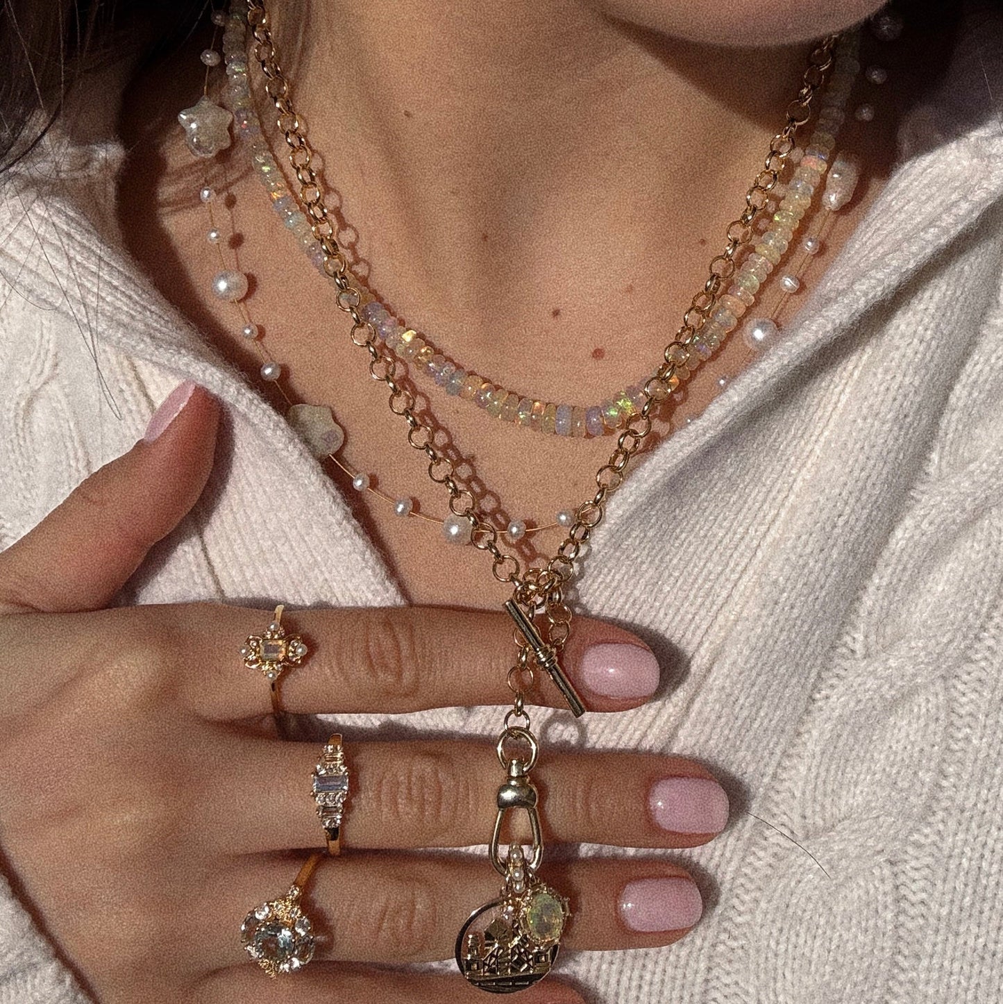A woman in a white knit sweater displays layered gold and pearl necklaces, including the Beaded Gemstone Necklace and an 18k gold plated piece. Her light pink manicured nails gently hold the necklaces, accenting her gemstone gold rings.