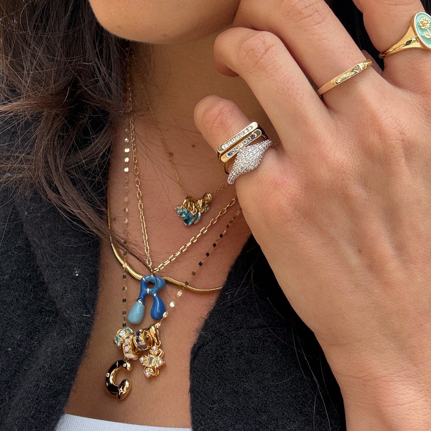 A close-up of a woman's hand with gold rings rests near her neck, layered with gold necklaces including the Star Crossed Celestial Bubble Letter Charm featuring sparkling jewels and blue, gold, and pearl accents. She wears a black top and coat.