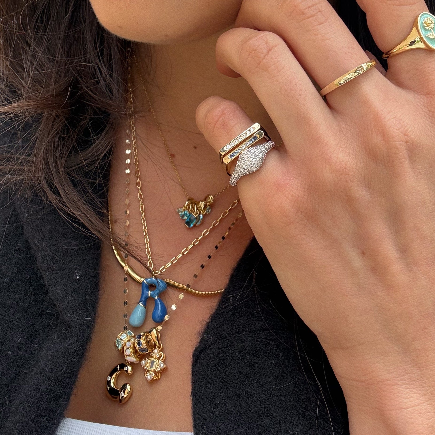 A close-up of a woman's hand with gold rings rests near her neck, layered with gold necklaces including the Star Crossed Celestial Bubble Letter Charm featuring sparkling jewels and blue, gold, and pearl accents. She wears a black top and coat.