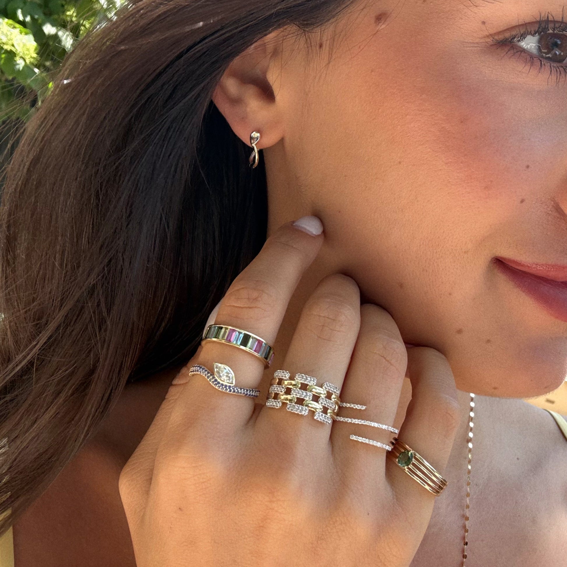 A woman with long brown hair wears multiple gold rings and the Solid Gold Mixed Metal Duo Huggies earring, holding her hand near her face to showcase Local Eclectic jewelry in the sunlight.