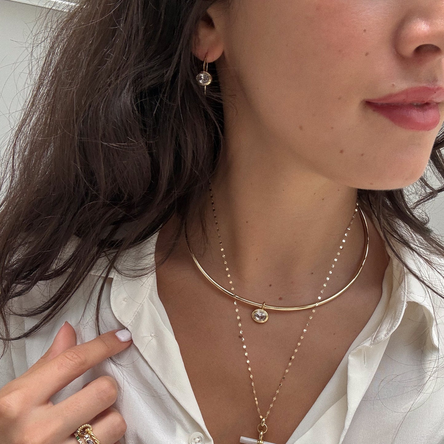 A woman in a white shirt showcases layered 18k yellow gold plated necklaces, a gold ring, and Lucia Oval French Hook Earrings. Her hand gently touches her collar, with her dark hair styled loosely.