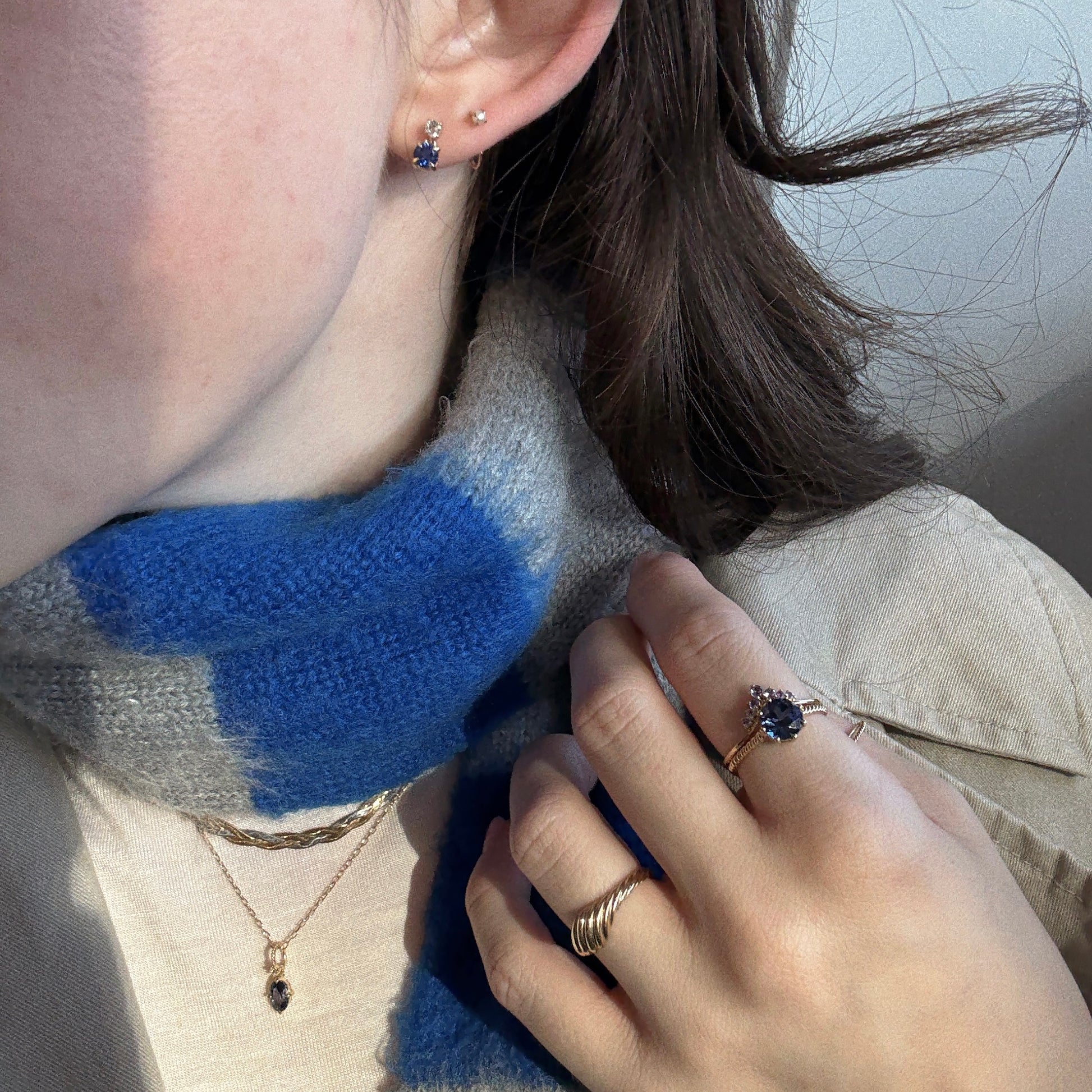A woman wearing a blue and gray scarf showcases the Solid Gold 2025 December Capsule Tanzanite Statement Set—earrings, necklace, and ring—plus a gold twisted ring, all against a neutral background.