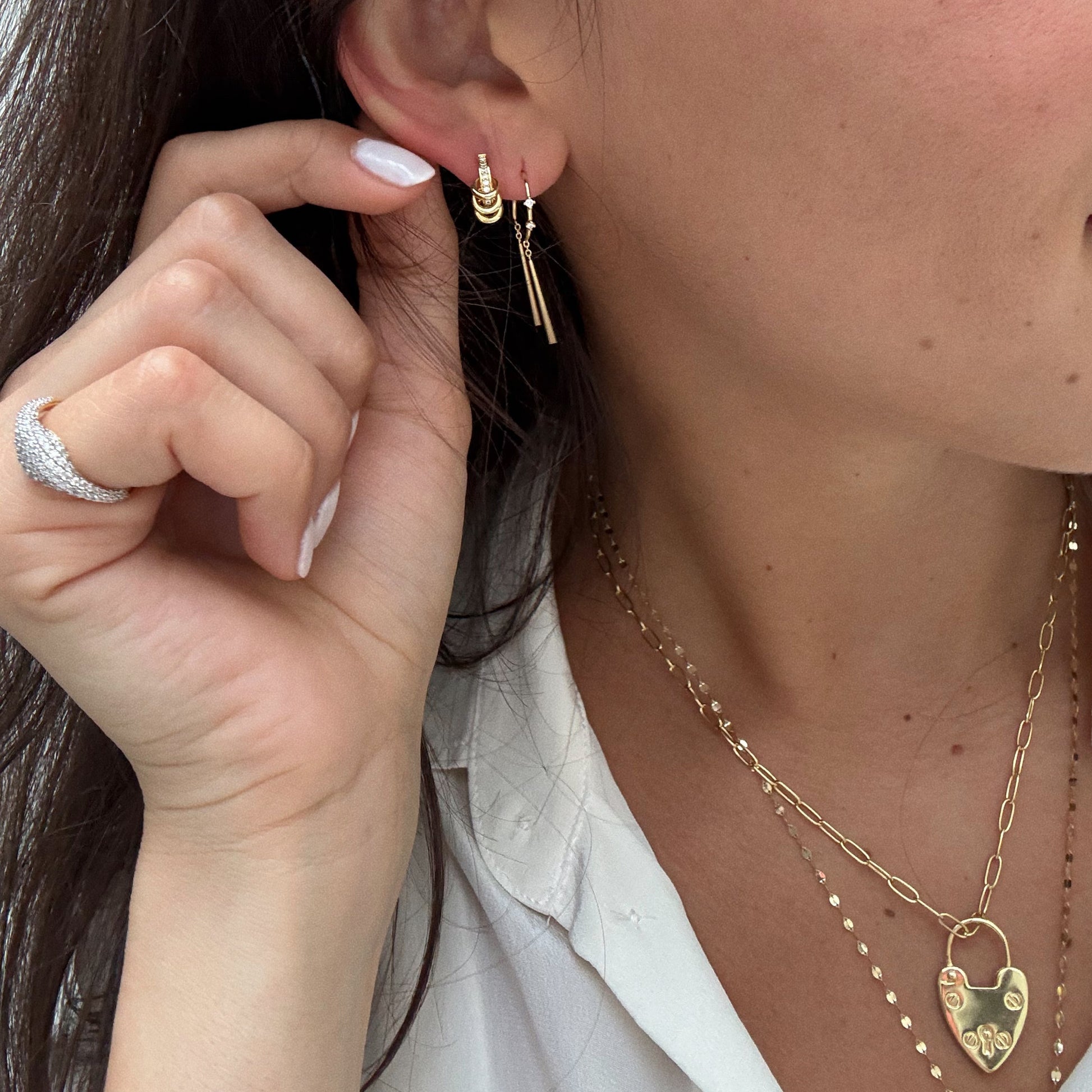 A woman in a white blouse wears layered 18k yellow gold plated necklaces, a heart-shaped pendant, Mila Threader Earrings, and a silver ring. She holds her ear to showcase her elegant jewelry collection.