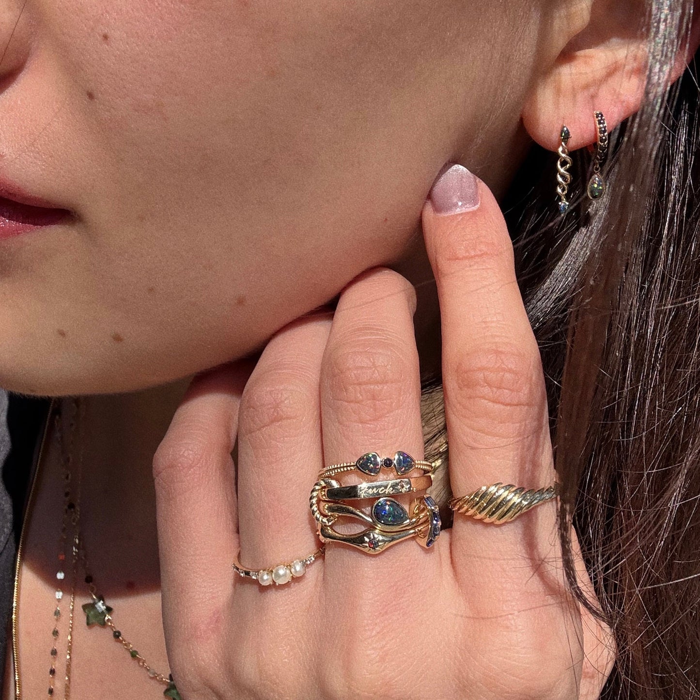 A close-up of a woman's face and hand shows gold rings with gemstones, a twisted gold ring, a pearl ring, and Solid Gold Black Opal Cosmic Spiral Earrings, as sunlight highlights her jewelry and skin.