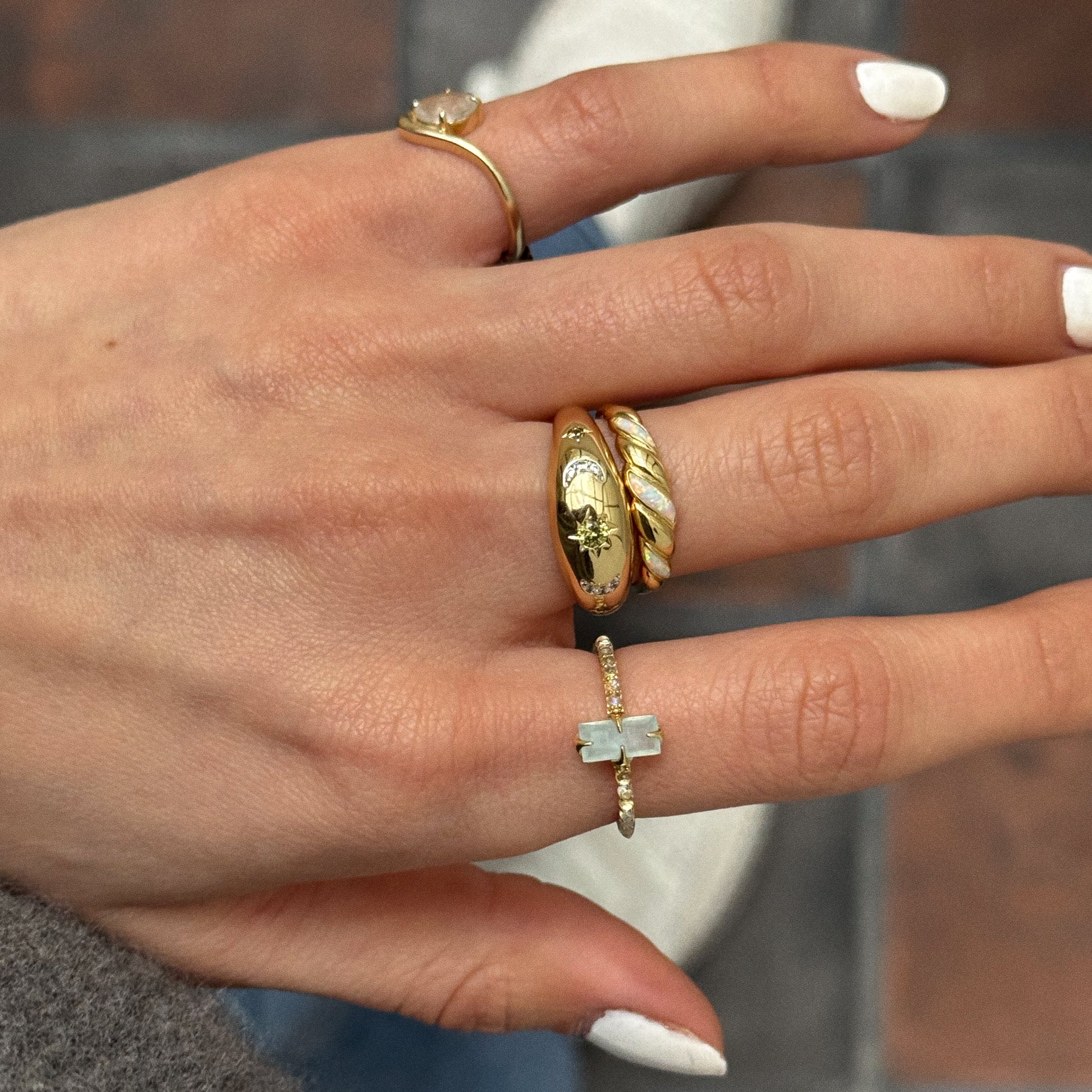 A hand with white nail polish wears four gold rings, one being the 2026 March Capsule Delicate Aquamarine Solitaire Ring, along with twisted, star, and cross designs. The background features a tiled floor.