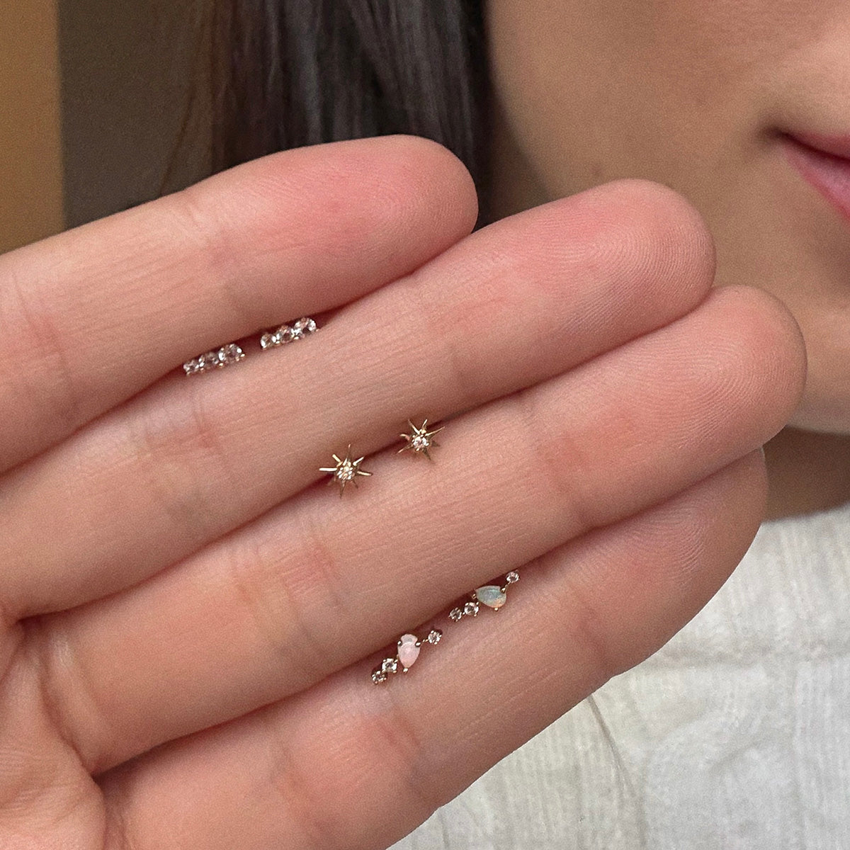 A close-up shows a hand holding two Solid Gold Starburst Flat Back Studs. The person wears several delicate solid gold earrings, with part of their face and white sweater visible in the background.
