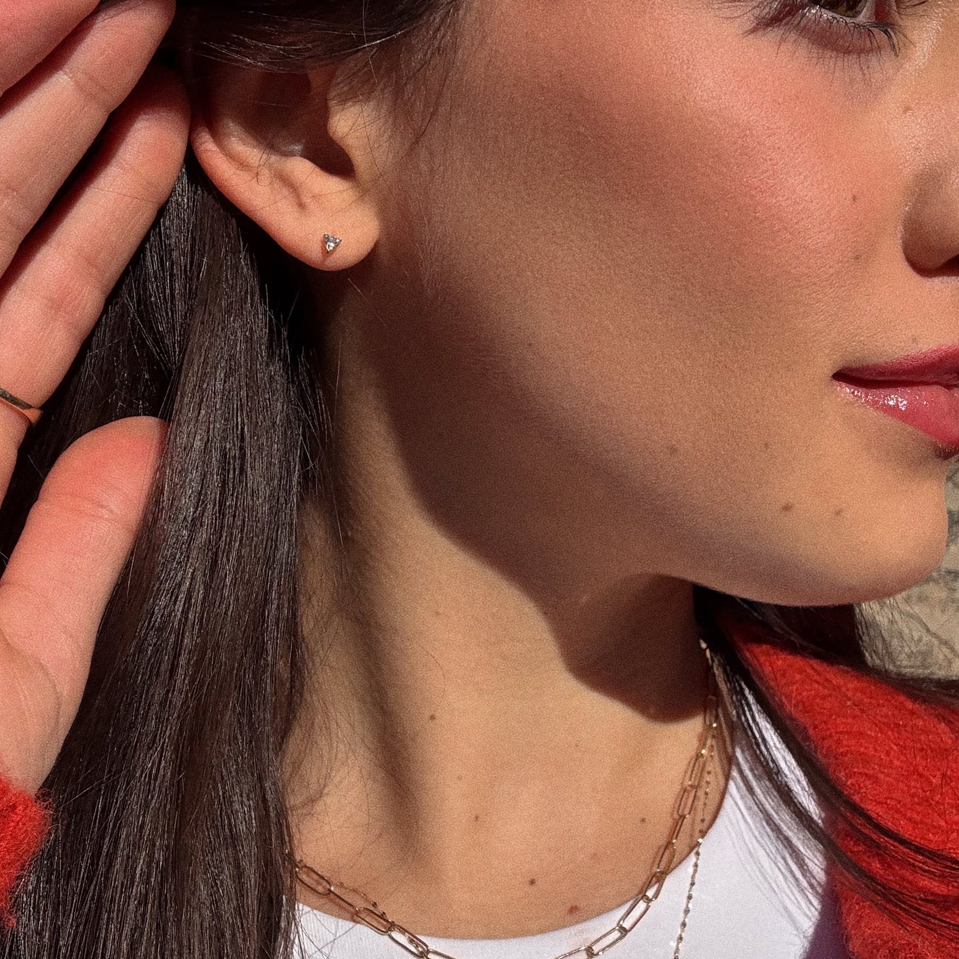 Close-up of a woman in sunlight with brown hair, glossy lips, and smooth skin, wearing a red top and gold necklace. She holds her hand near her ear, which is adorned with Solid Gold White Topaz You Have My Heart Studs.