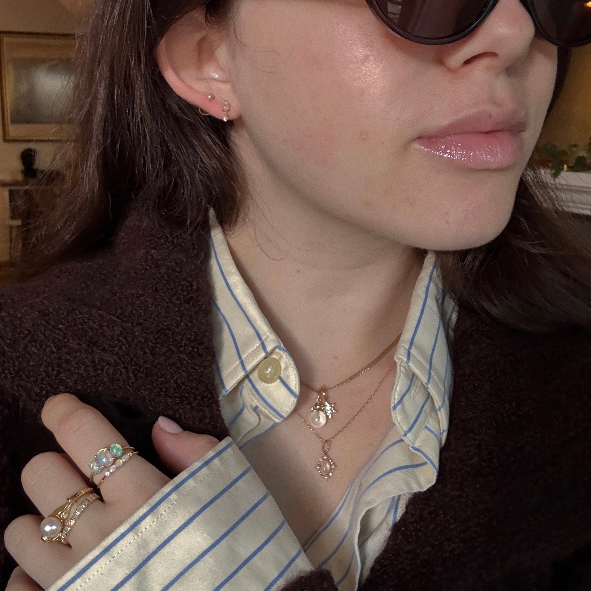 A person with glossy lips and sunglasses wears a striped shirt, brown coat, pearl necklace, and the Solid Gold 2025 October Capsule Opal Statement Set. A softly lit background showcases framed artwork. Free gift included.