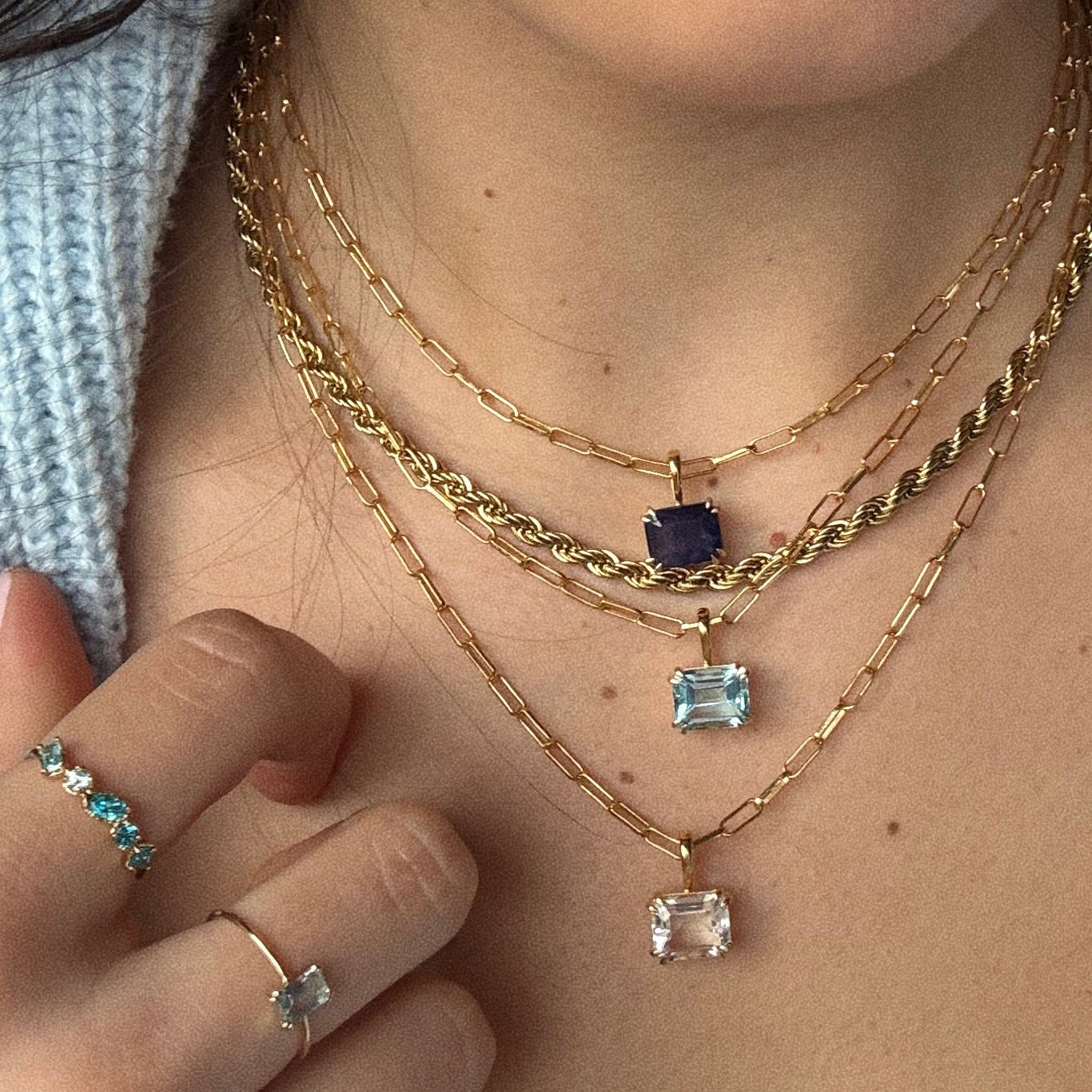 A woman wears Born to Shine Birthstone Necklaces with square blue, light blue, and pale pink pendants and gold rings featuring blue and clear stones, paired with a light grey knit sweater.