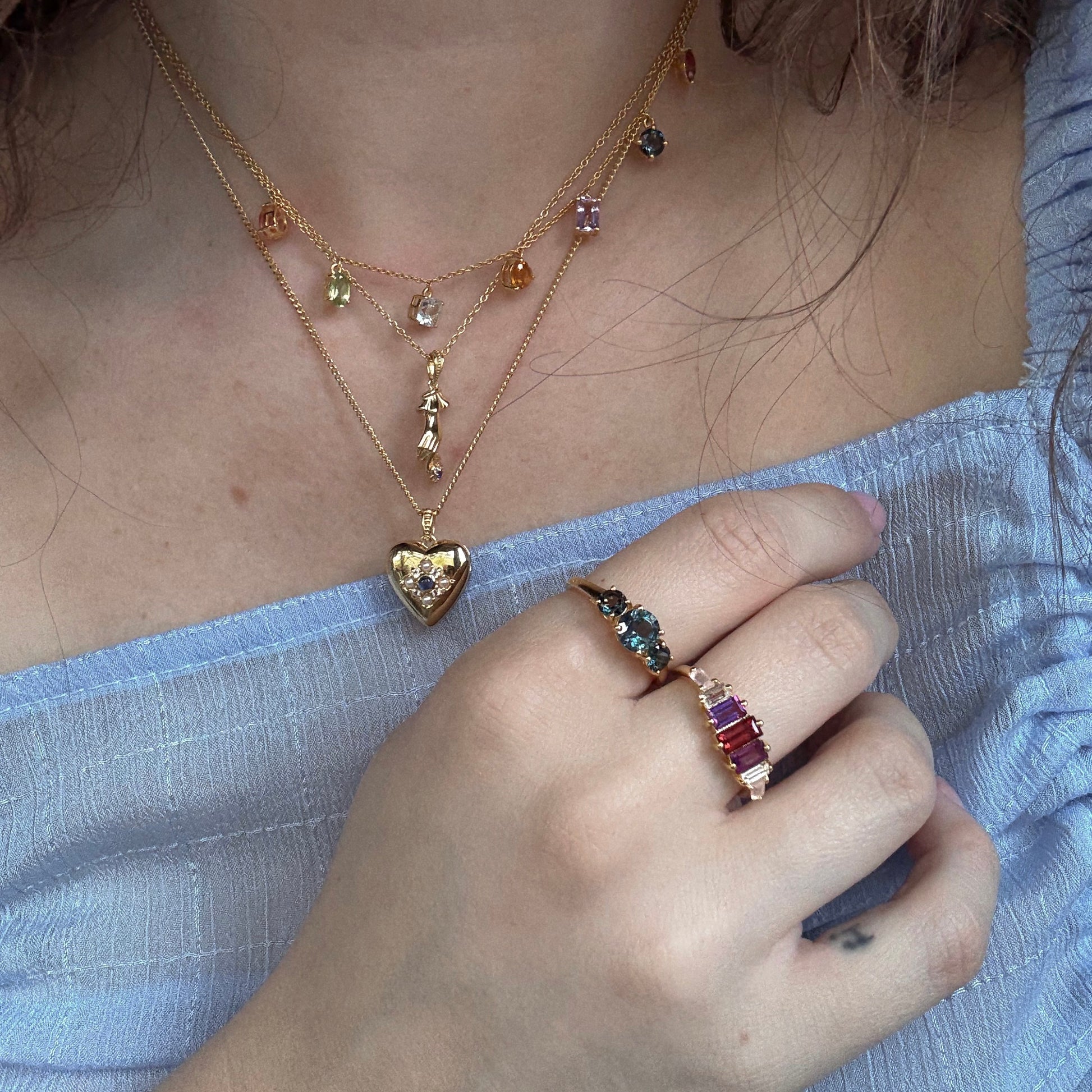 A person wears layered 14k gold vermeil necklaces, one with a heart-shaped pendant, and the Berry Baroque Garnet Crown Ring. They are dressed in a light blue, off-shoulder, textured top.