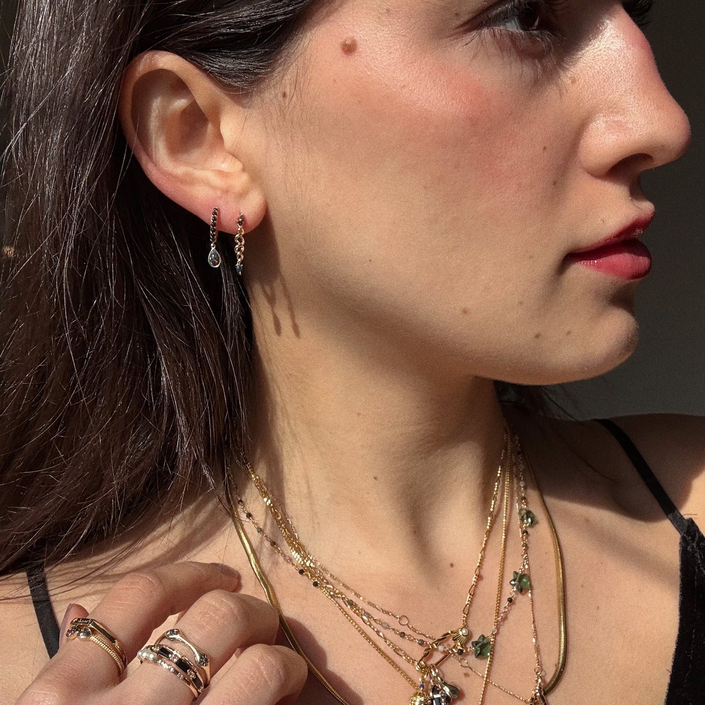 A woman with long brown hair and light skin, dressed in a black top, is shown in profile wearing Solid Gold Black Opal Cosmic Spiral Earrings, along with layered gold necklaces and rings. Sunlight casts shadows on her face.