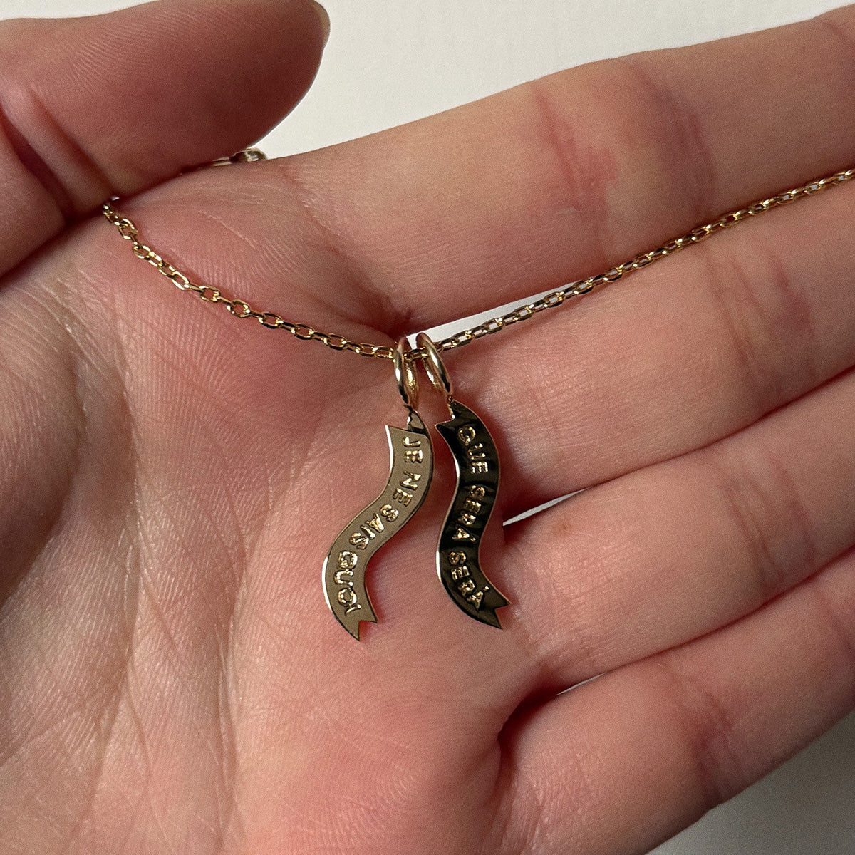 A hand holds Charms by I Like It Here Club, a gold chain necklace with 12k yellow gold plating and two wavy charms engraved with SEE THE GOOD and SEE THE BEAUTY, against a light background that highlights its personalized flair.