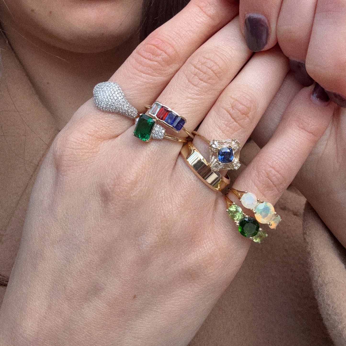 Two manicured hands display multiple gold rings, including the Solid Gold Aquamarine Sapphire and Garnet Baguette Gemstone Ring (Sample Size 9), with colorful gemstones like green, blue, red, and opal against a neutral background.