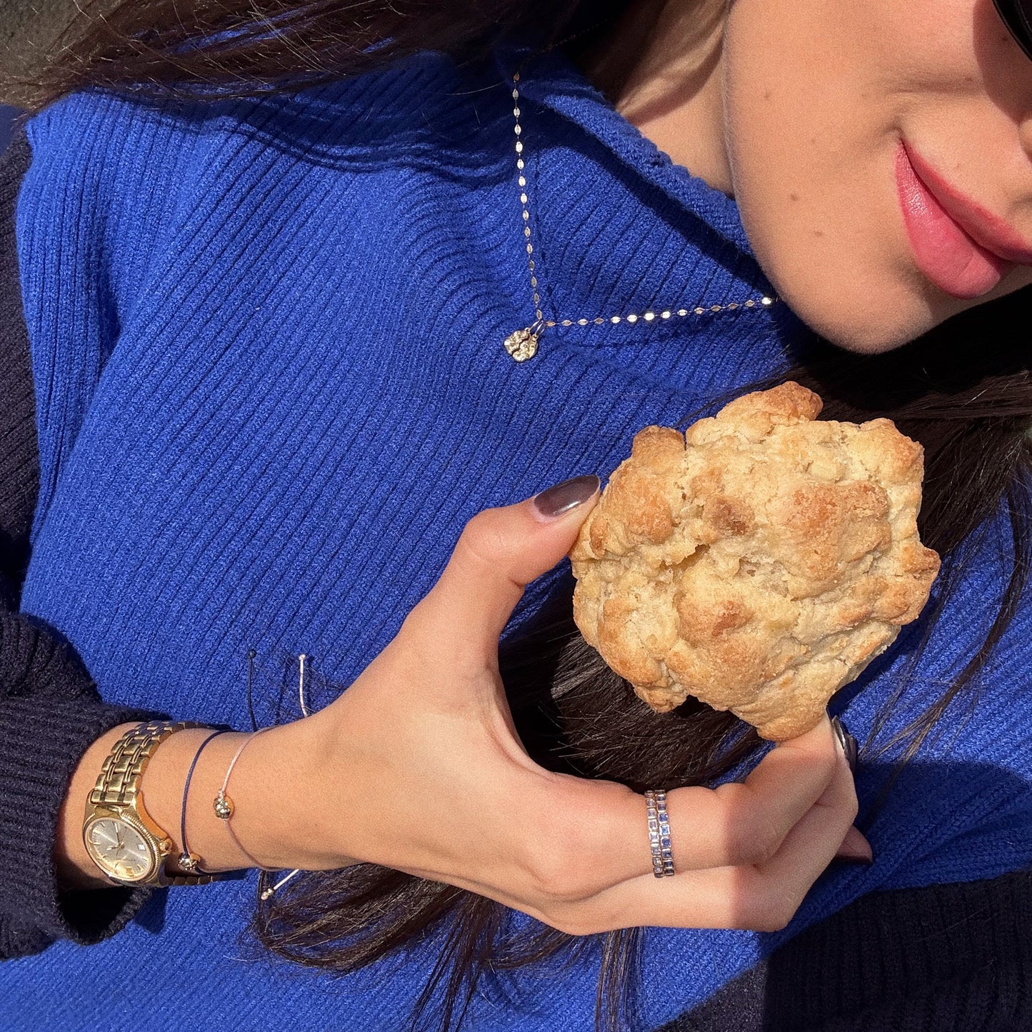 A woman with long brown hair, pink lipstick, sunglasses, and painted nails holds a large rustic cookie while wearing the Levain Bakery x Local Eclectic Solid Gold Share a Cookie Friendship Charm with her blue sweater and gold accessories.