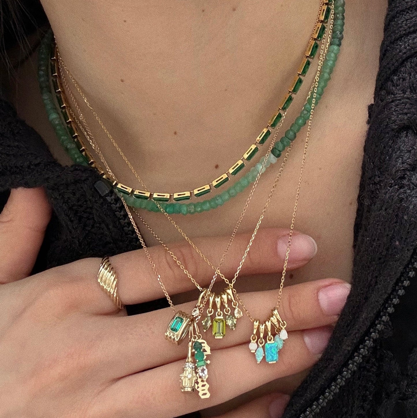 A person wearing the Shades of You Ombre Birthstone Charm Necklace with green, blue, and clear gemstone charms, a green beaded 16" necklace, and a gold twisted ring, dressed in a dark textured top.