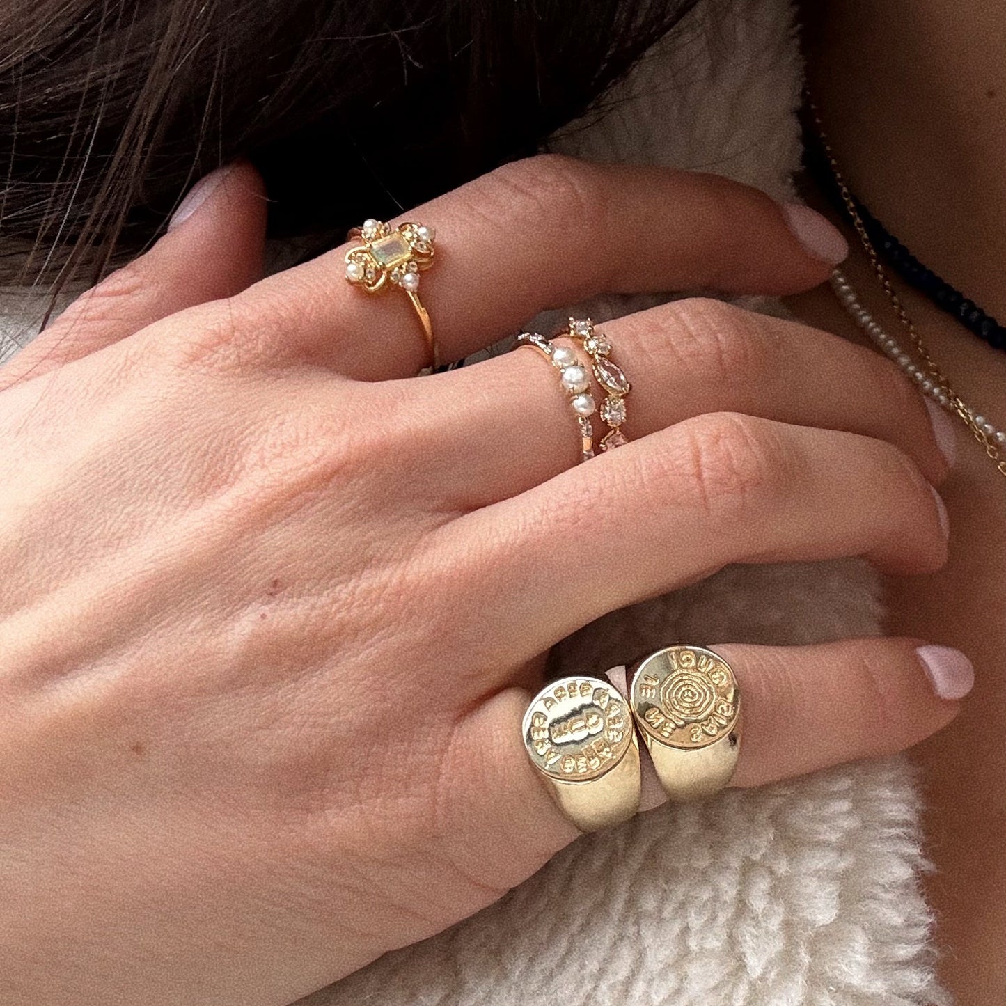 A close-up of a hand adorned with multiple rings, including the I Like It Here Club x Local Eclectic Je Ne Sais Quoi Pinky Ring, gold signet rings, and delicate stone-studded bands. The person is dressed in a cream textured garment.