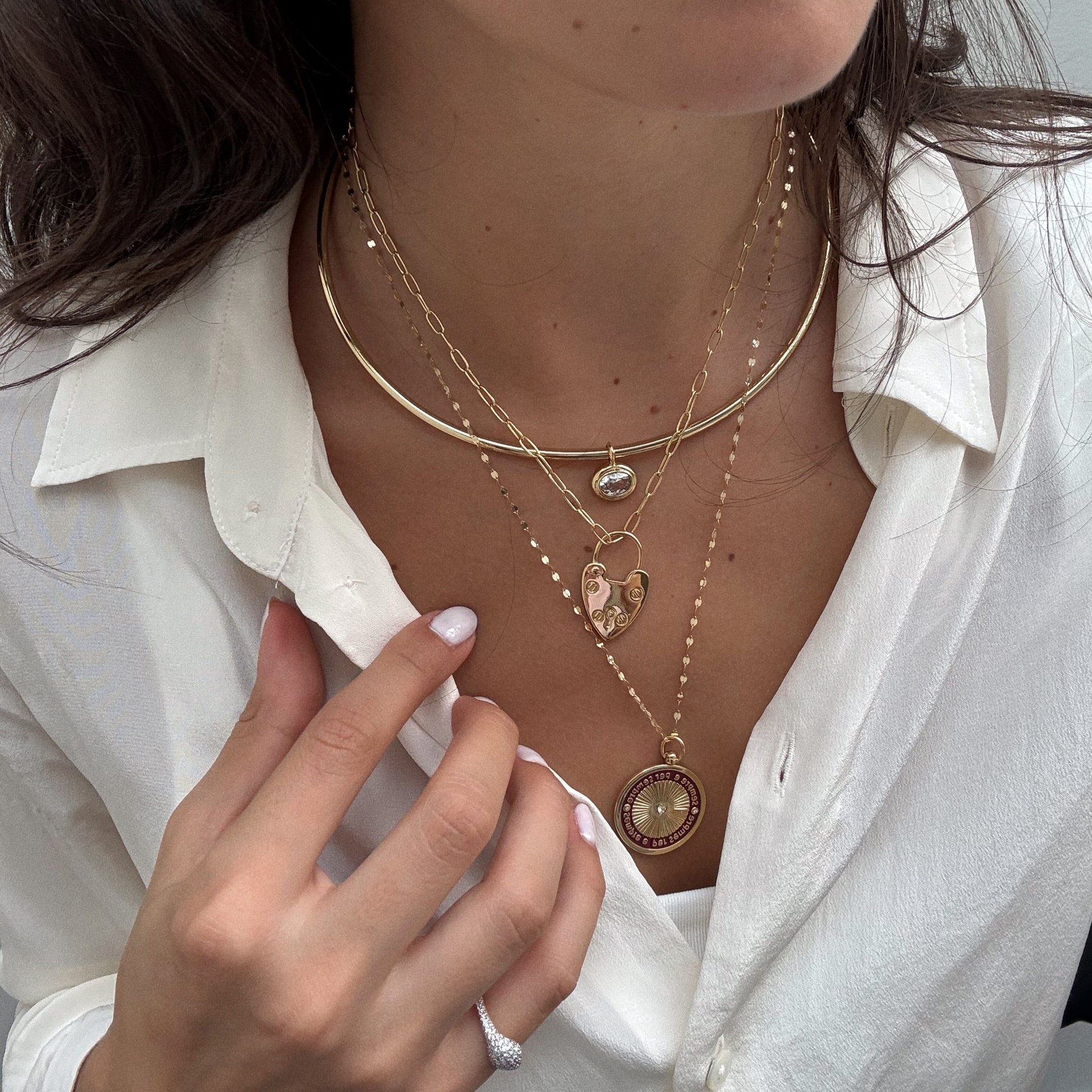 A woman in a white button-up shirt models four layered gold necklaces, featuring the Hayes Collar Necklace with heart, coin, and circular pendants. She holds one necklace and wears a ring on her finger.