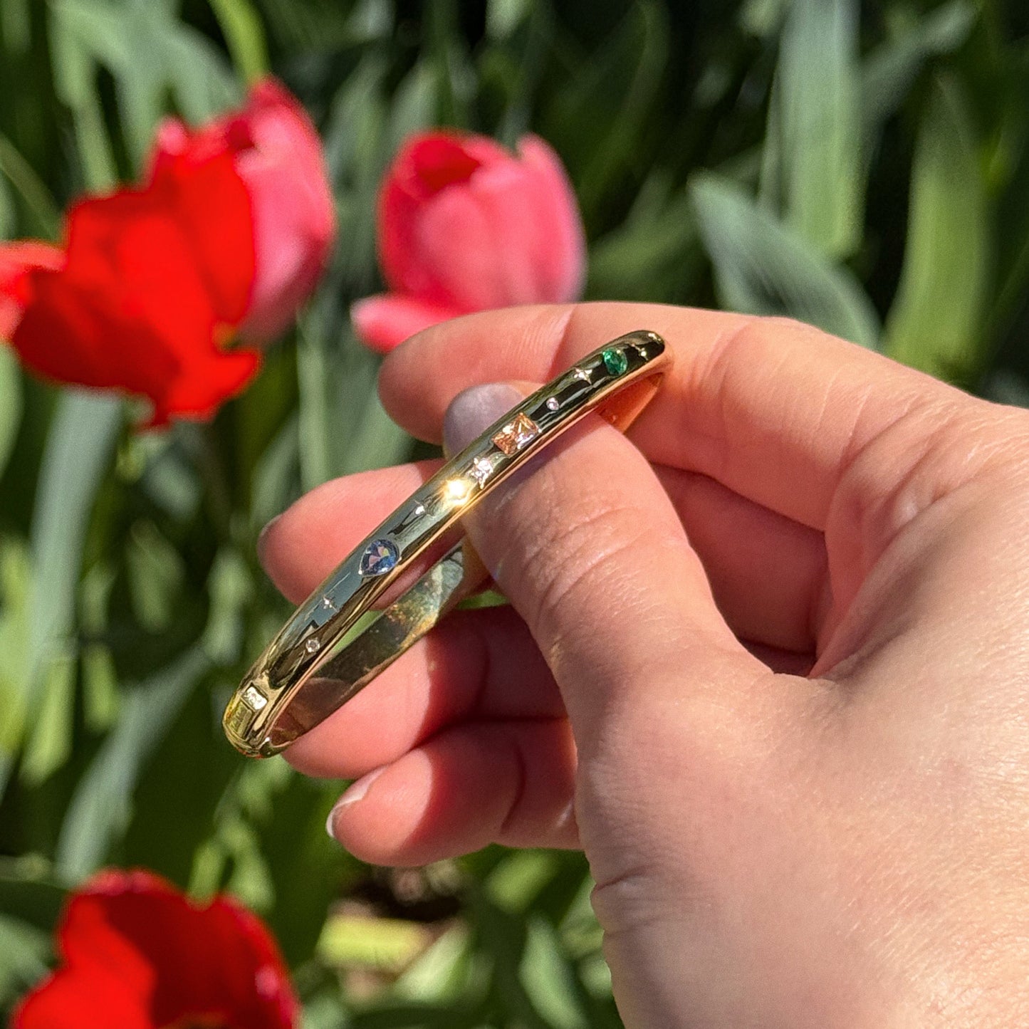 A hand holds the Hidden Curse Word Fuckin A Bracelet, a Local Eclectic exclusive gold plated bangle with colorful gemstones, in bright sunlight with red tulips and green foliage blurred in the background.