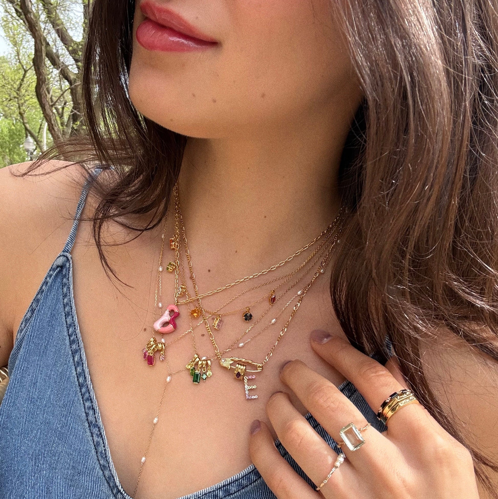 A woman in a blue denim top wears layered gold necklaces, featuring the Solid Gold Pearl Lariat Necklace with freshwater pearls. She sports stacked rings and wavy brown hair, set against a backdrop of trees.
